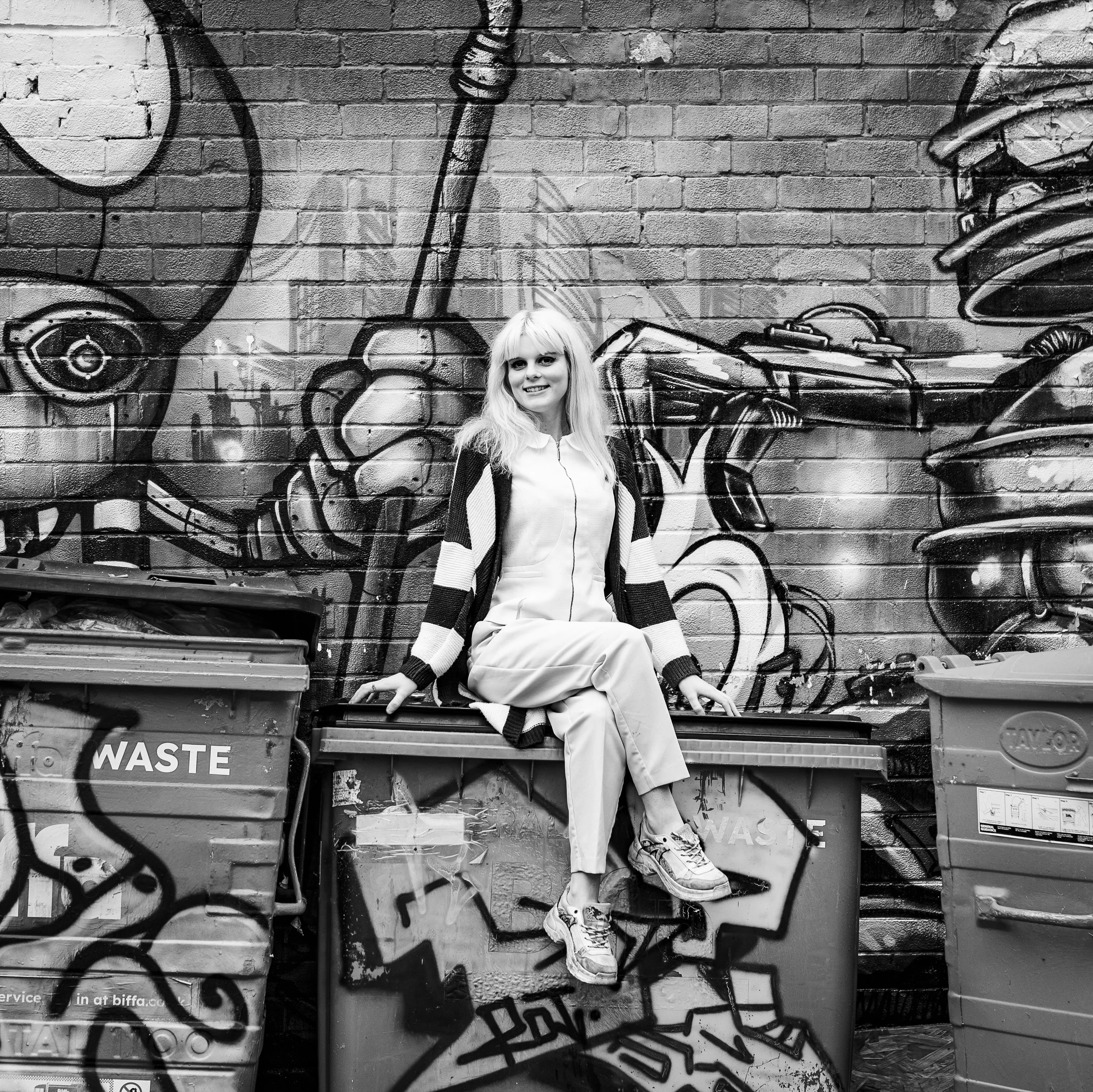 A woman sitting on a graffiti-covered dumpster in front of a brick wall with graffiti art, in black and white.