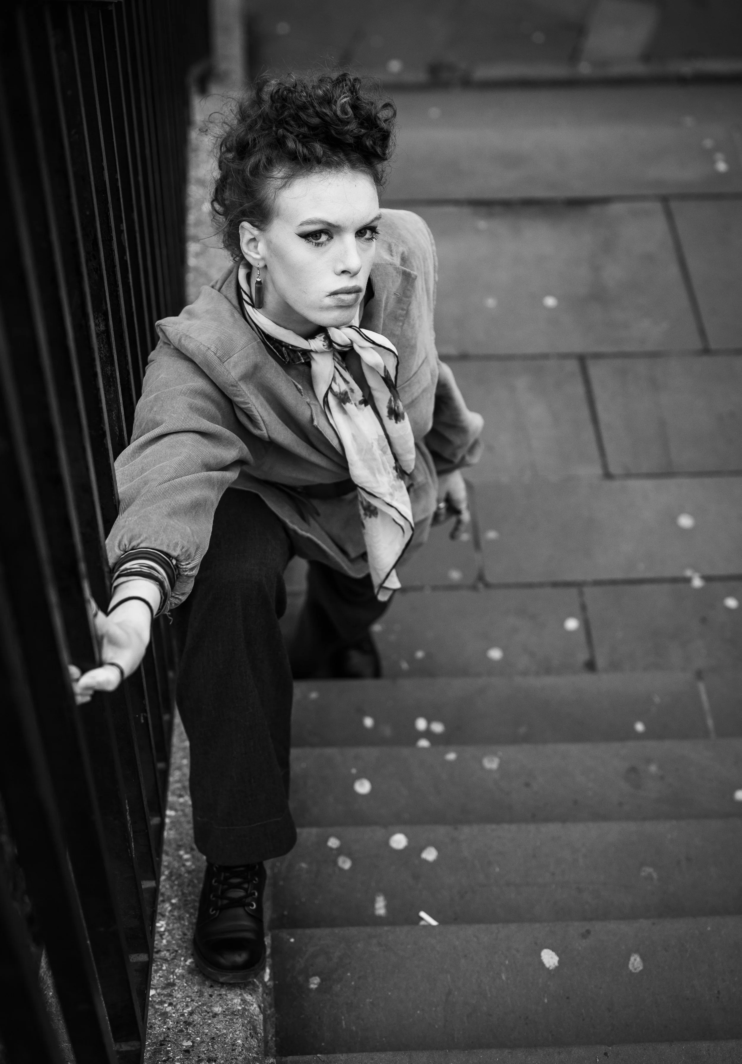 A woman with curly hair and makeup looking up at the camera while sitting on outdoor stairs, holding onto a railing, wearing a jacket, scarf, and dark pants in black and white.