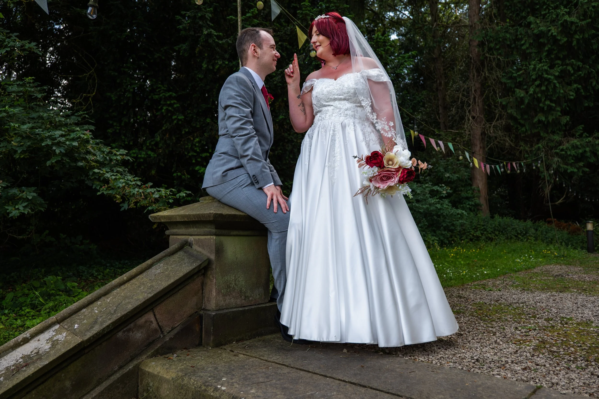 A bride with red hair wearing a white wedding gown and veil holding a bouquet of red, pink, and white flowers, standing in front of a groom with short hair in a gray suit, sitting on a stone ledge outdoors surrounded by greenery, with festive bunting