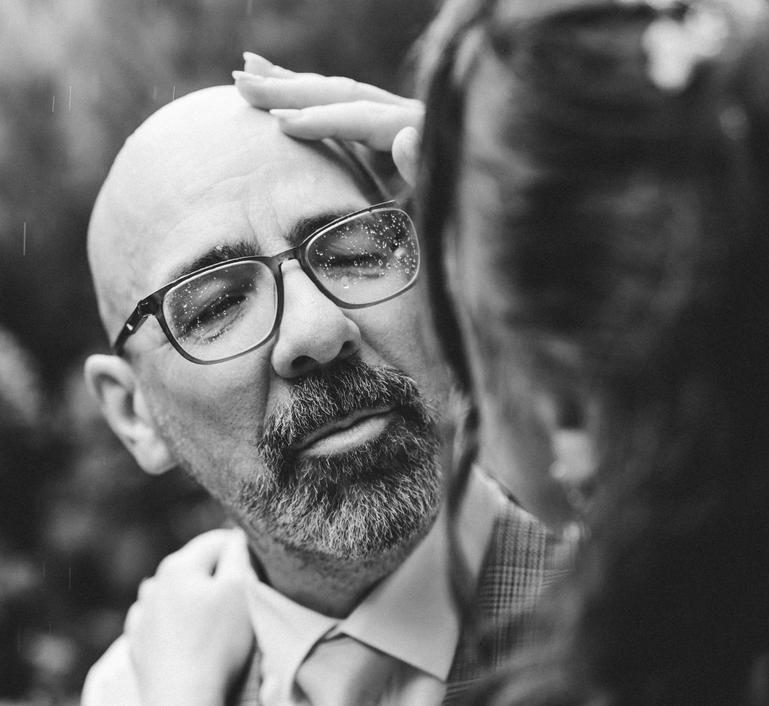 Black and white close-up of a man with glasses, a beard, and a mustache, touching his forehead with one hand.
