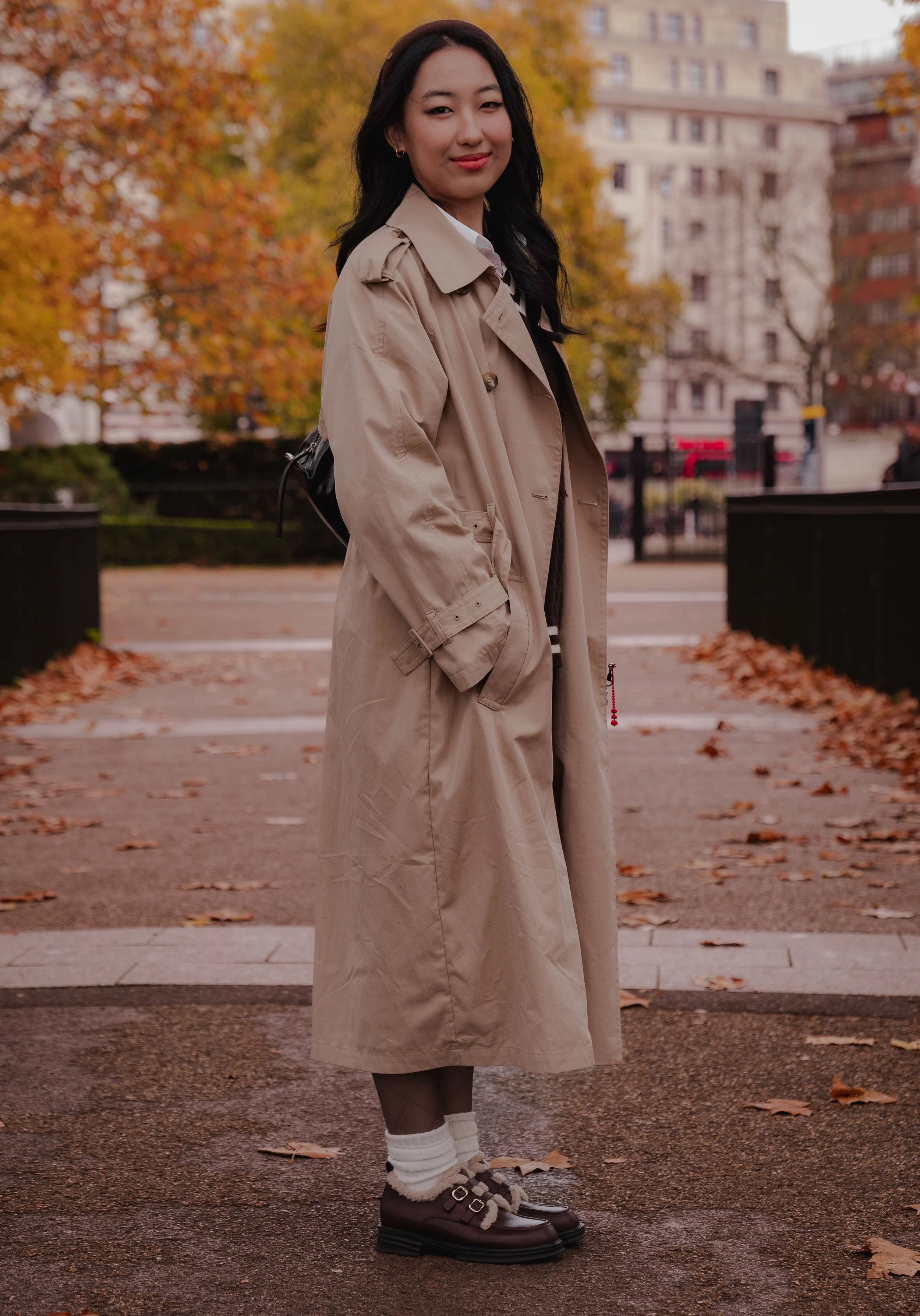 A woman in a beige trench coat standing on a fall sidewalk with autumn leaves and trees in the background, smiling at the camera.