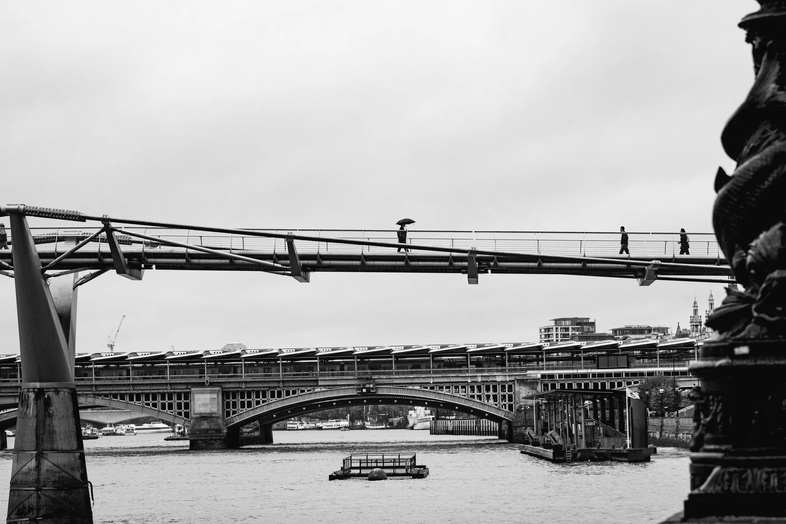 Black and white photo of a bridge over a river, with three pedestrians walking across; one person is holding an umbrella. Urban buildings and a cloudy sky in the background.