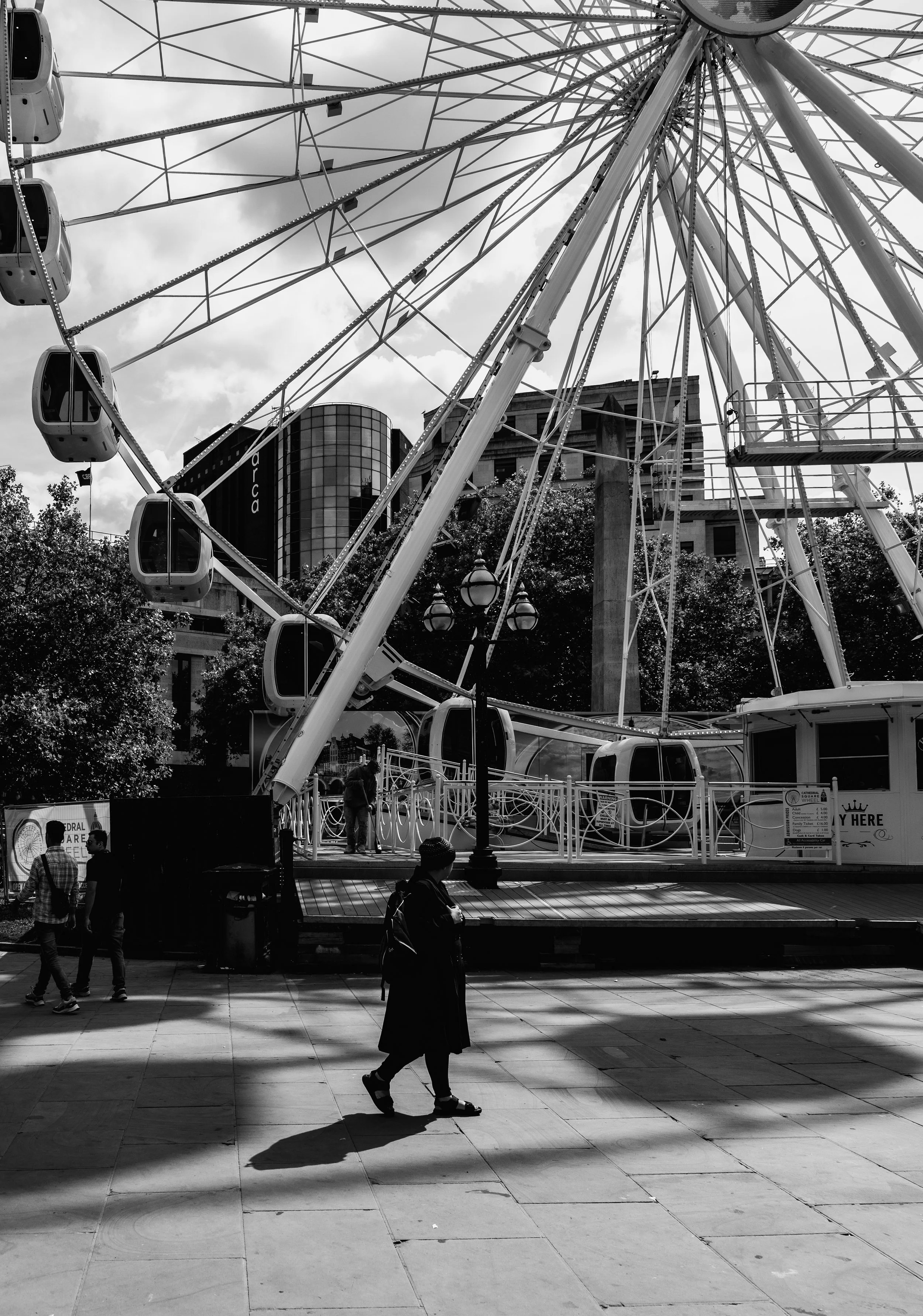 A black and white photo of a large outdoor Ferris wheel with enclosed gondolas, set against a cityscape with modern buildings and trees, with a person walking on the sidewalk in the foreground.
