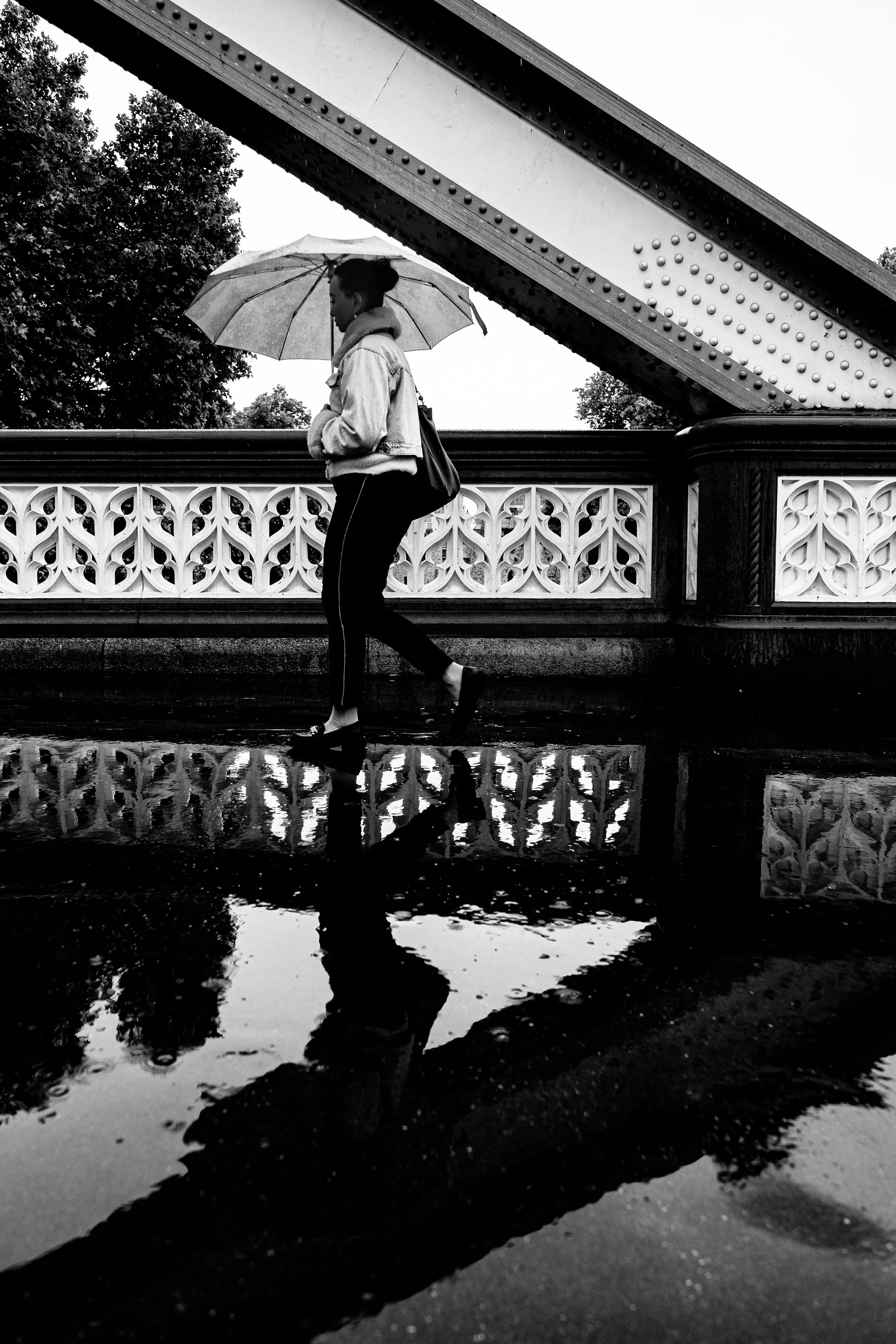 A woman walking with an umbrella on a rainy day, reflected in a puddle, near a bridge with ornate railings.