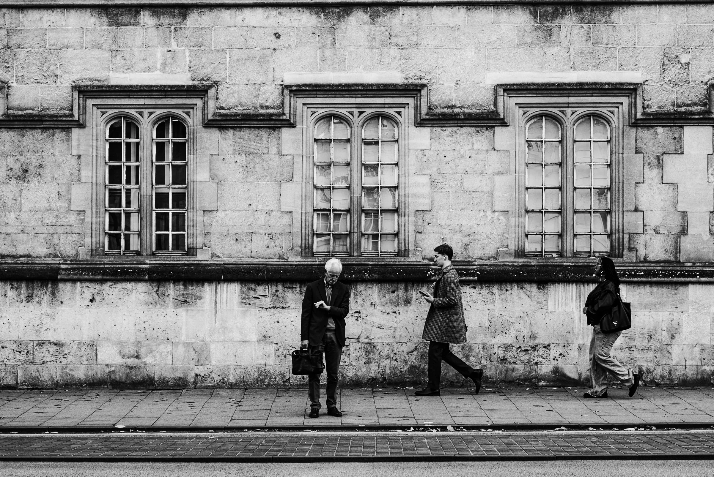 Three people walking on a sidewalk against a stone building with three arched windows, two men and a woman in business attire, all using their phones.