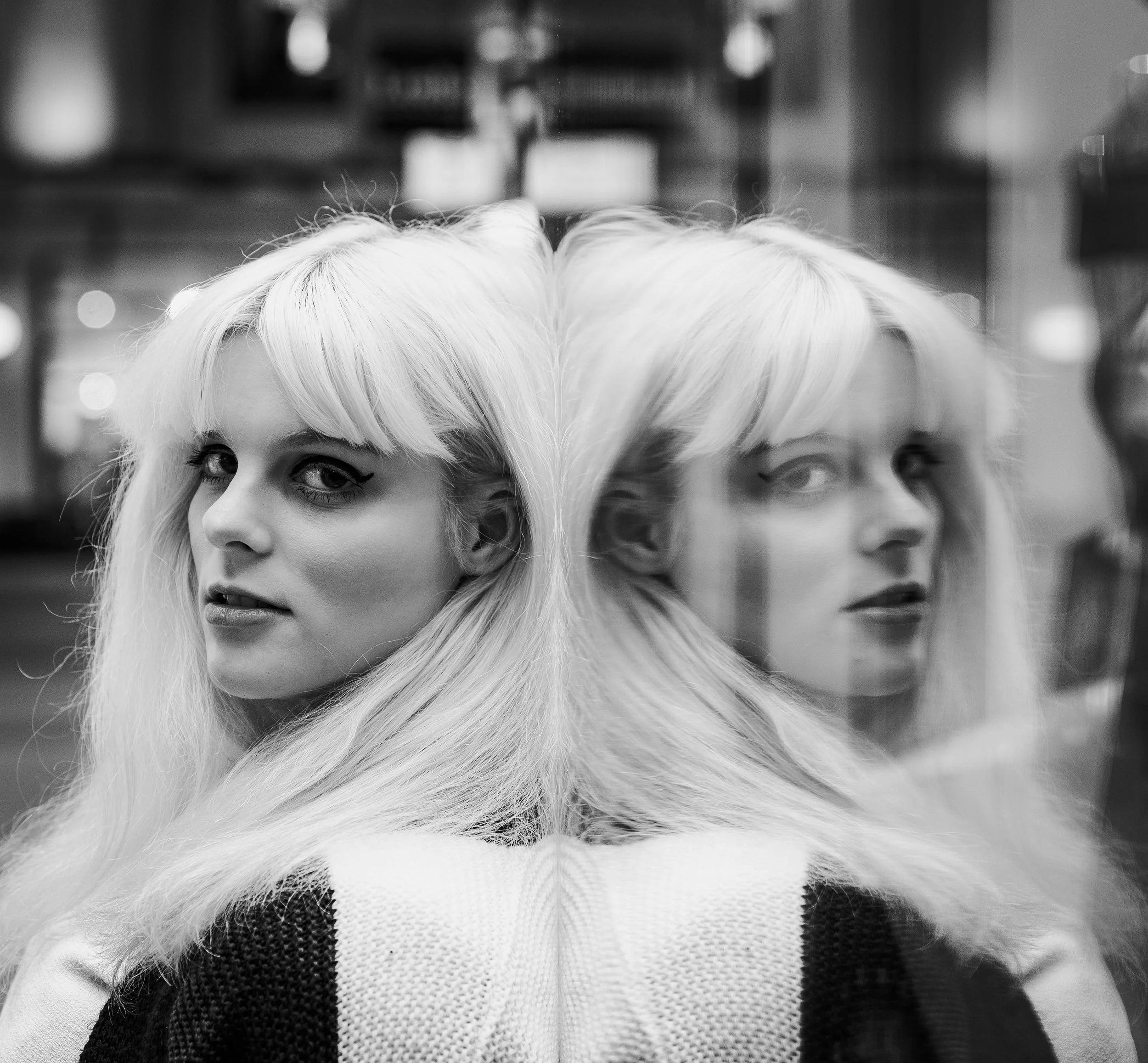 Black and white photo of a woman with light-colored hair looking at the camera, with her reflection visible on a glass surface beside her.