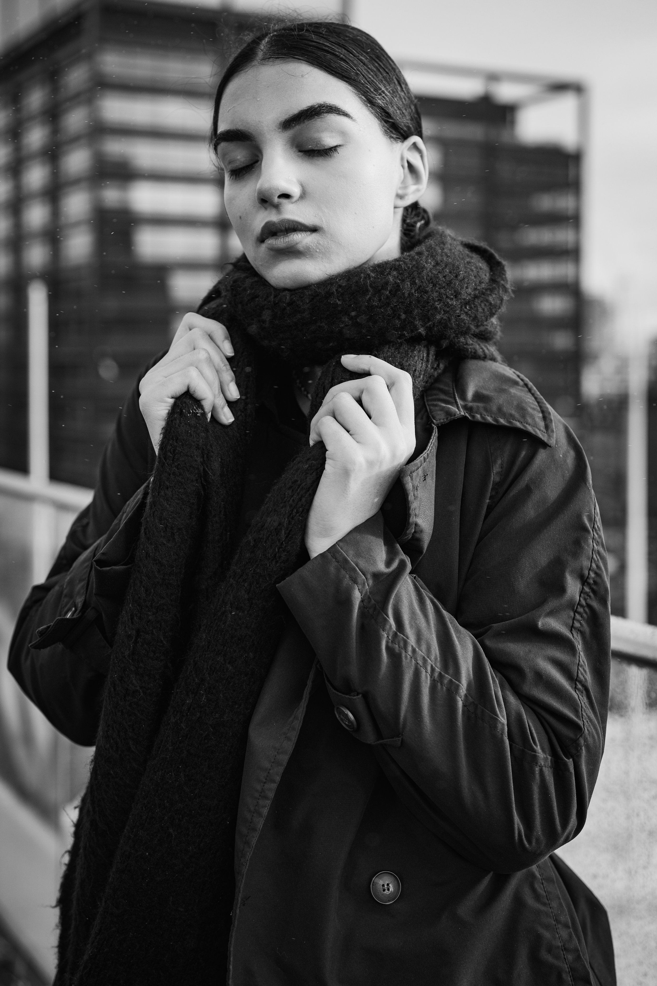 A black and white photograph of a woman with closed eyes, holding a scarf or coat around her neck. She is wearing a coat and appears to be outdoors in an urban setting.