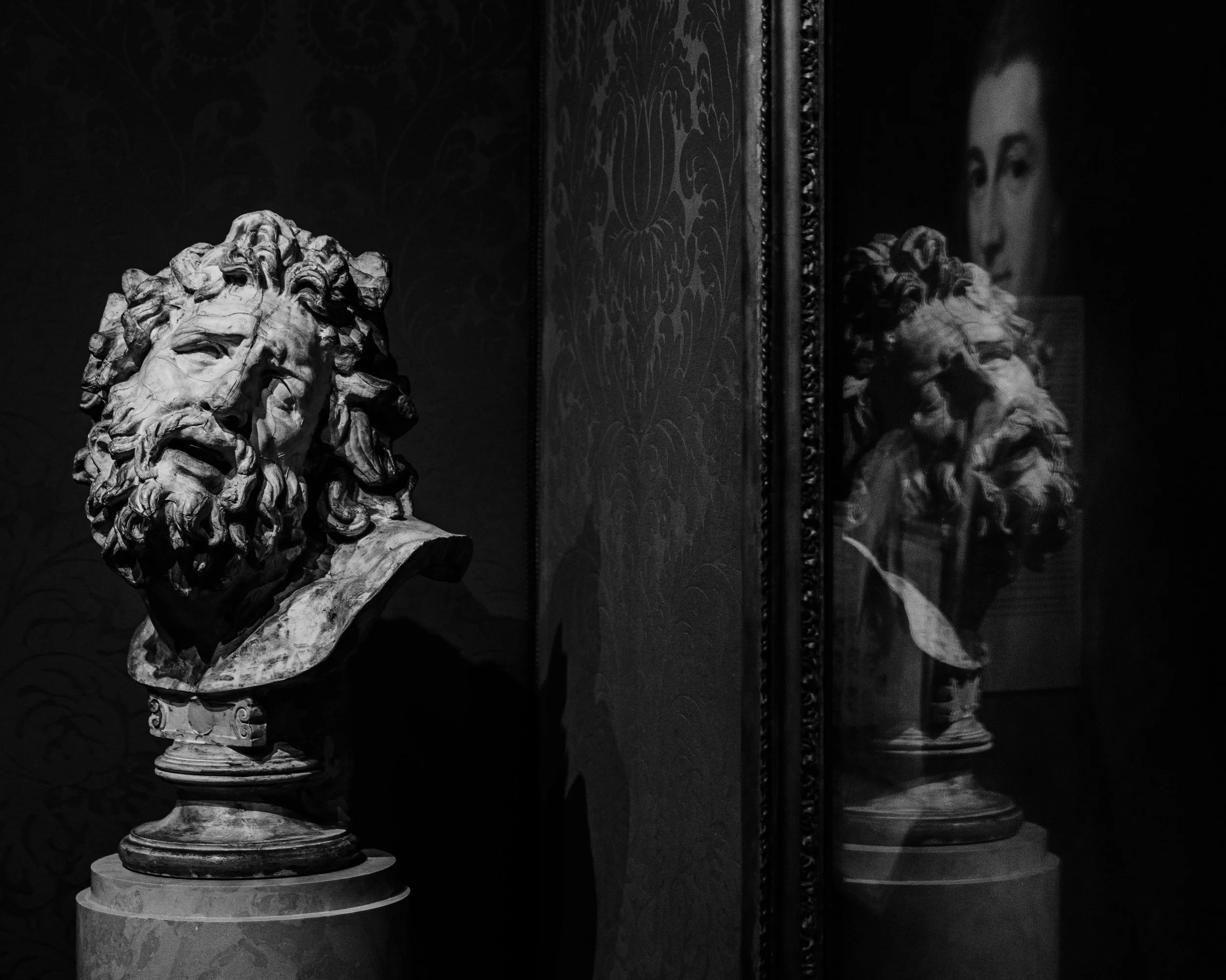 A marble sculpture of a bearded man with long, curly hair displayed in a shadowed museum setting. The sculpture's reflection is visible in a framed mirror on the right.
