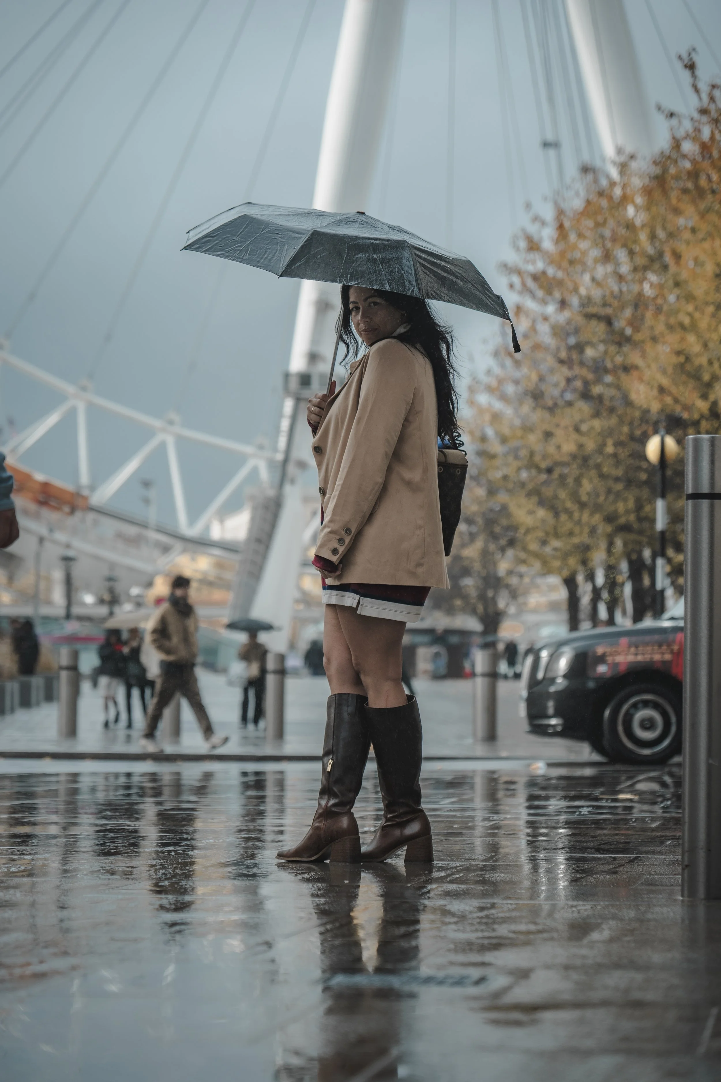 A woman standing on a wet city street holding an umbrella, wearing a beige coat, shorts, and knee-high boots, with a large Ferris wheel in the background and autumn trees.