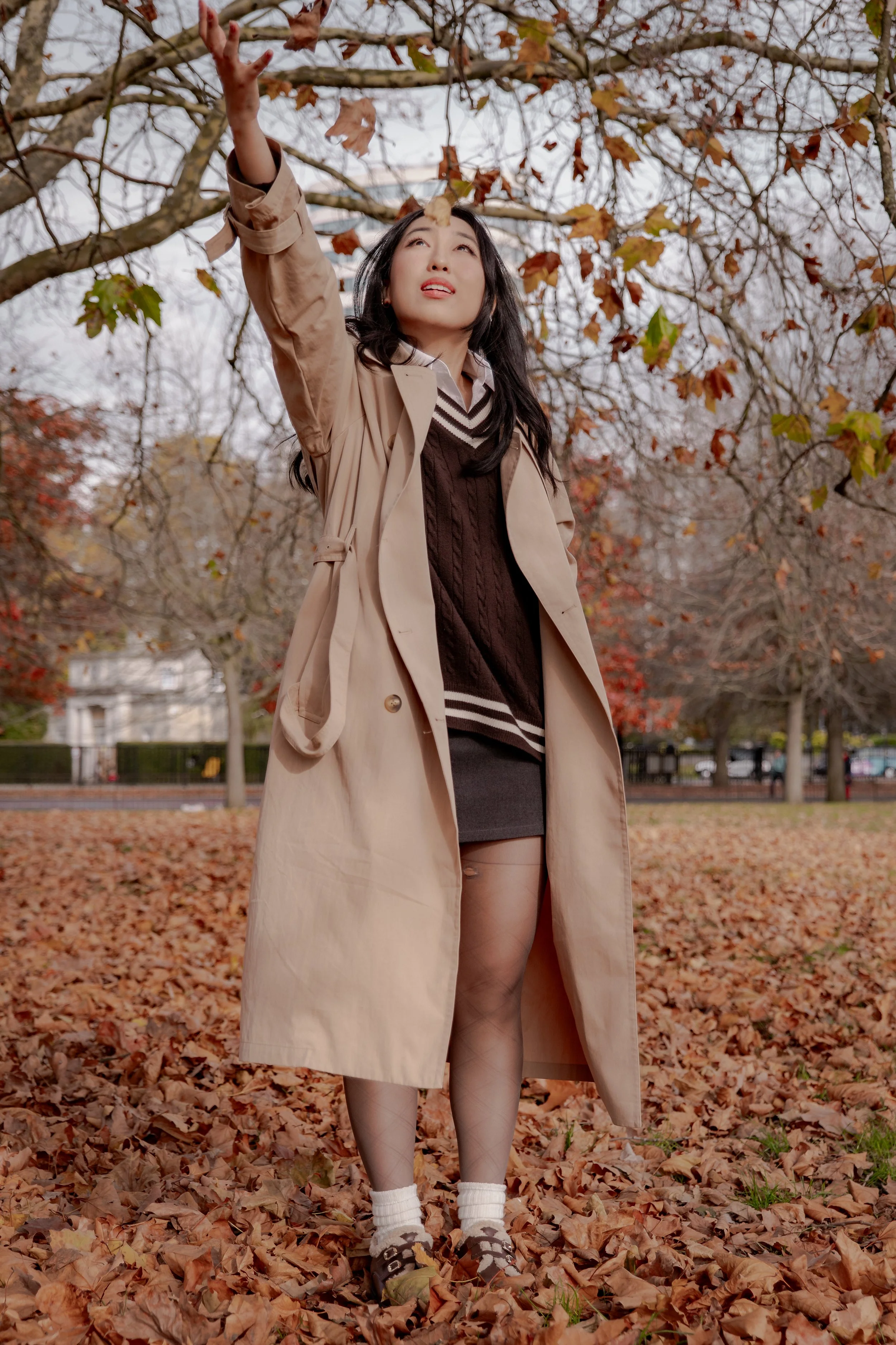 A woman in a tan trench coat and black sweater standing in a park covered in fallen autumn leaves, reaching up towards a tree branch with colorful leaves.