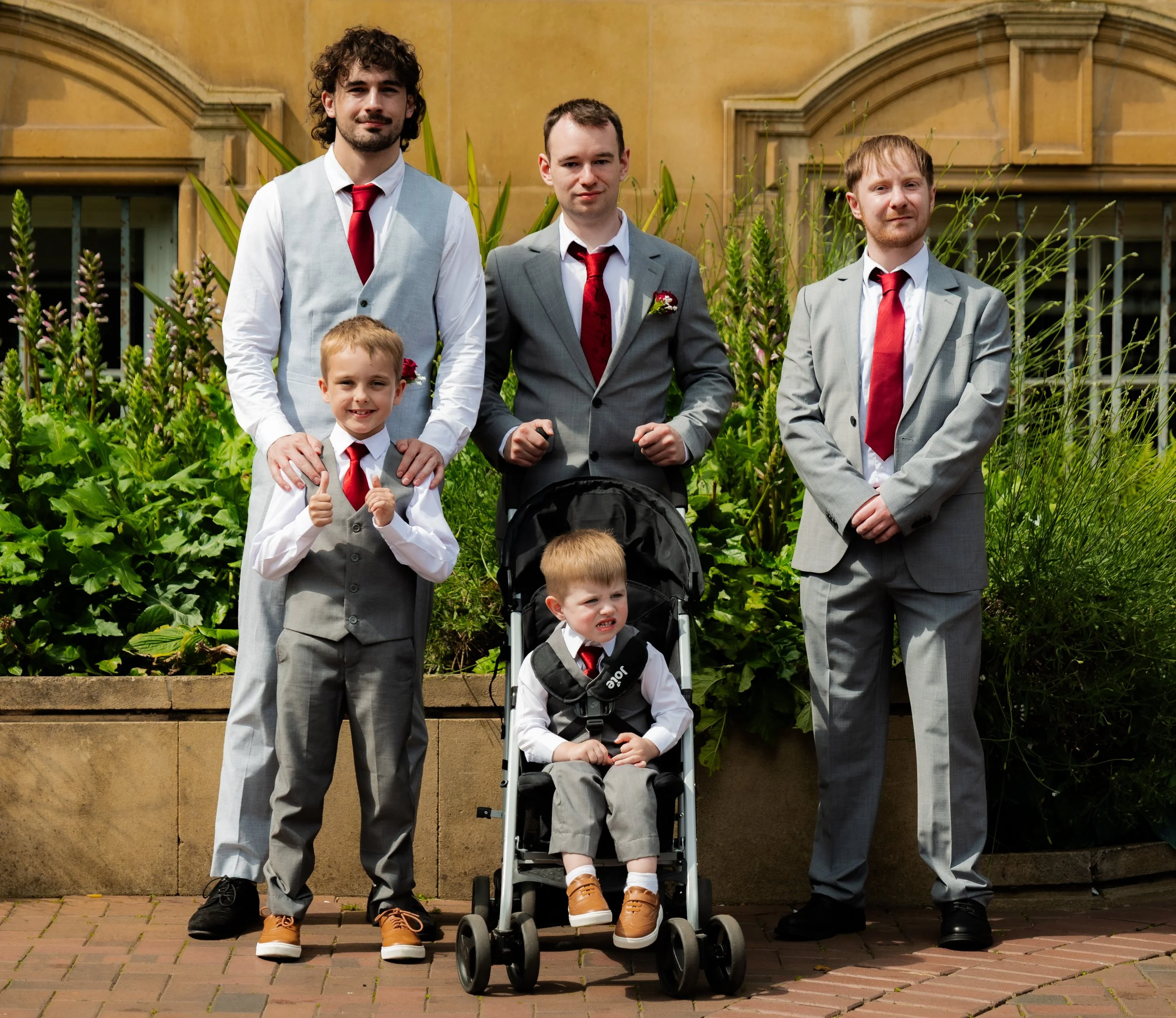 Seven males, including one young boy in a stroller, dressed in formal suits with red ties, posing outdoors with greenery and a building in the background.