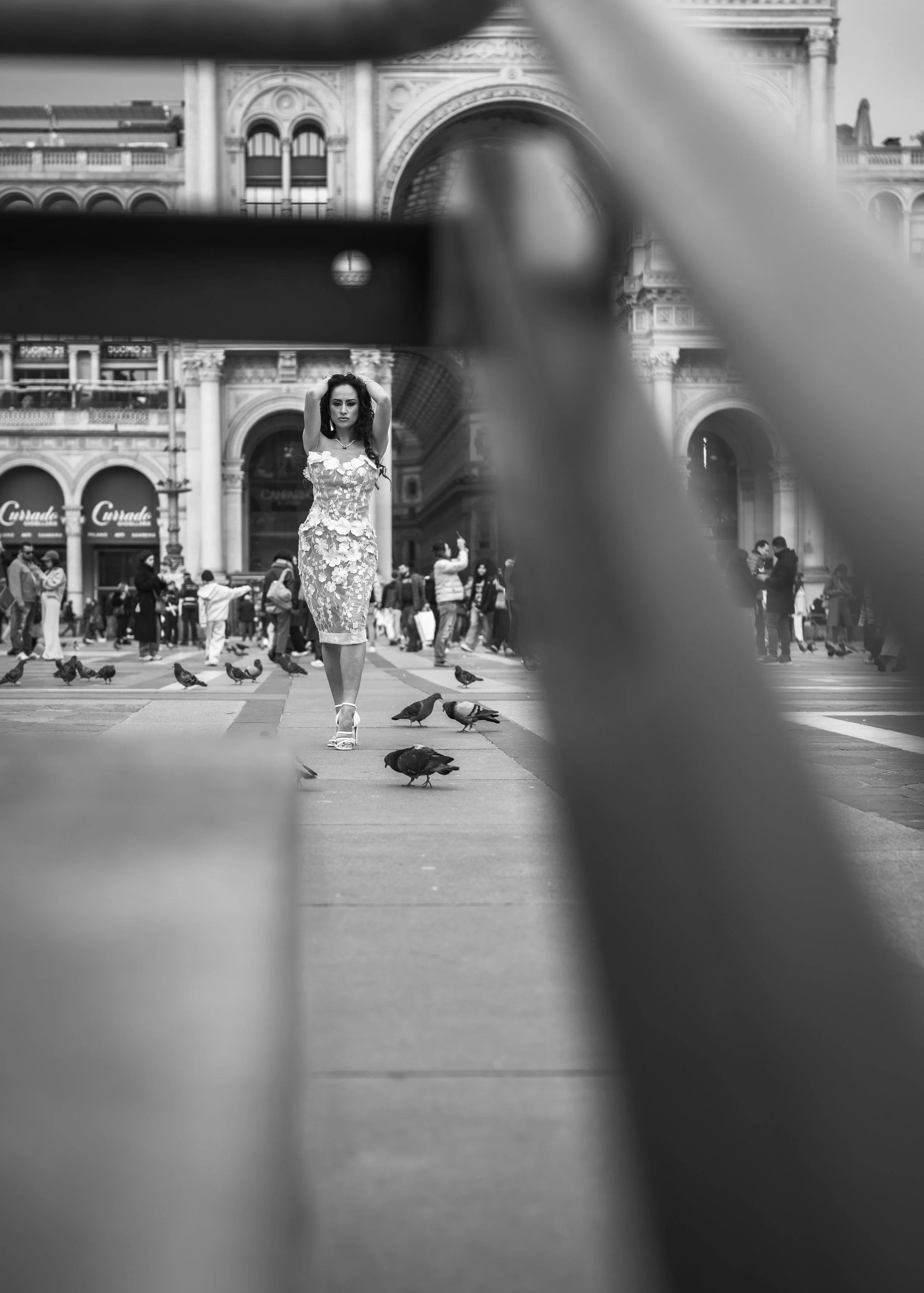 A woman in a lace dress walking through a busy city square with pigeons, seen through a metal structure.
