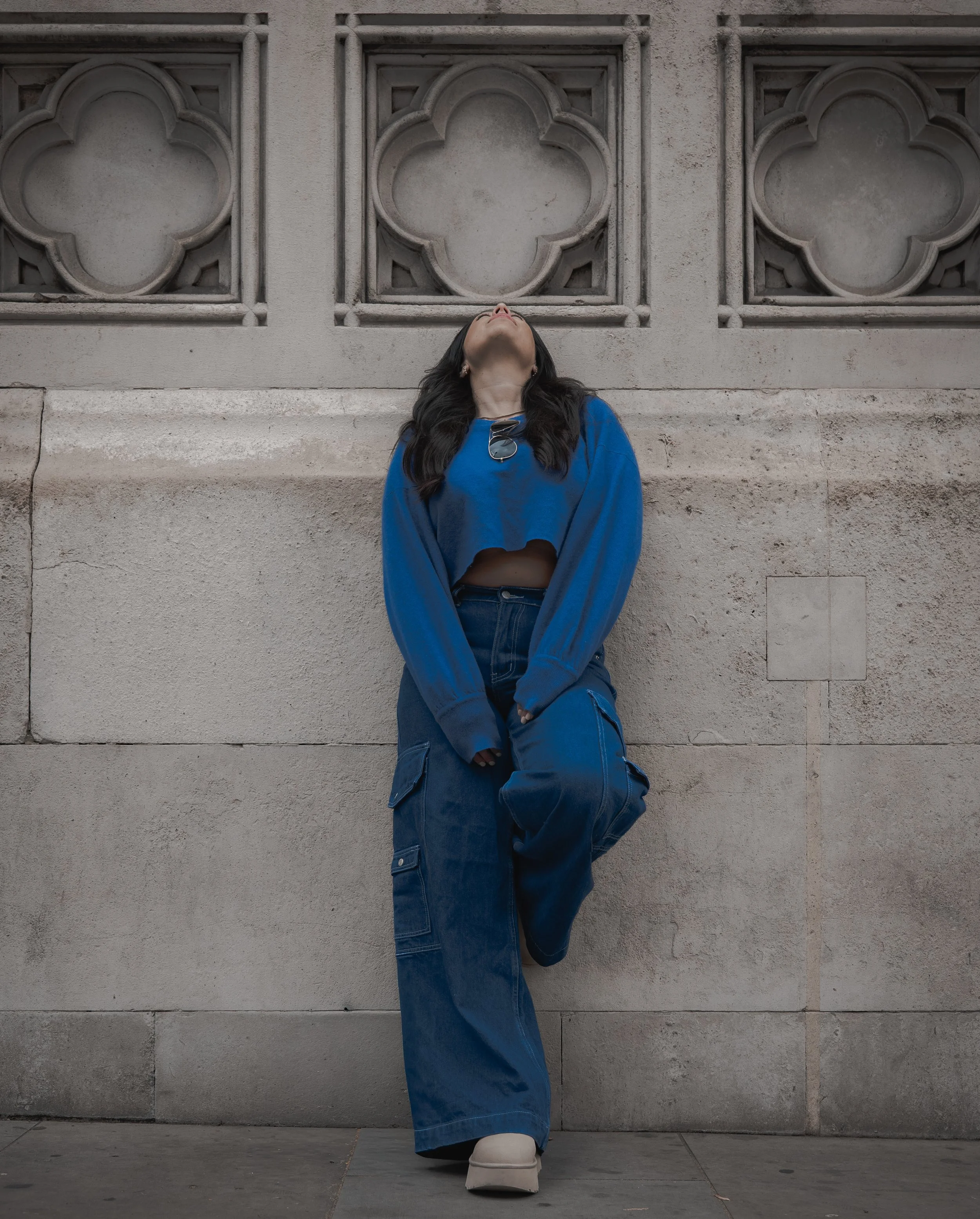 A woman in a blue sweatshirt, cargo pants, and white shoes standing against a textured stone wall with decorative window openings, looking upwards with sunglasses hanging from her shirt neckline.