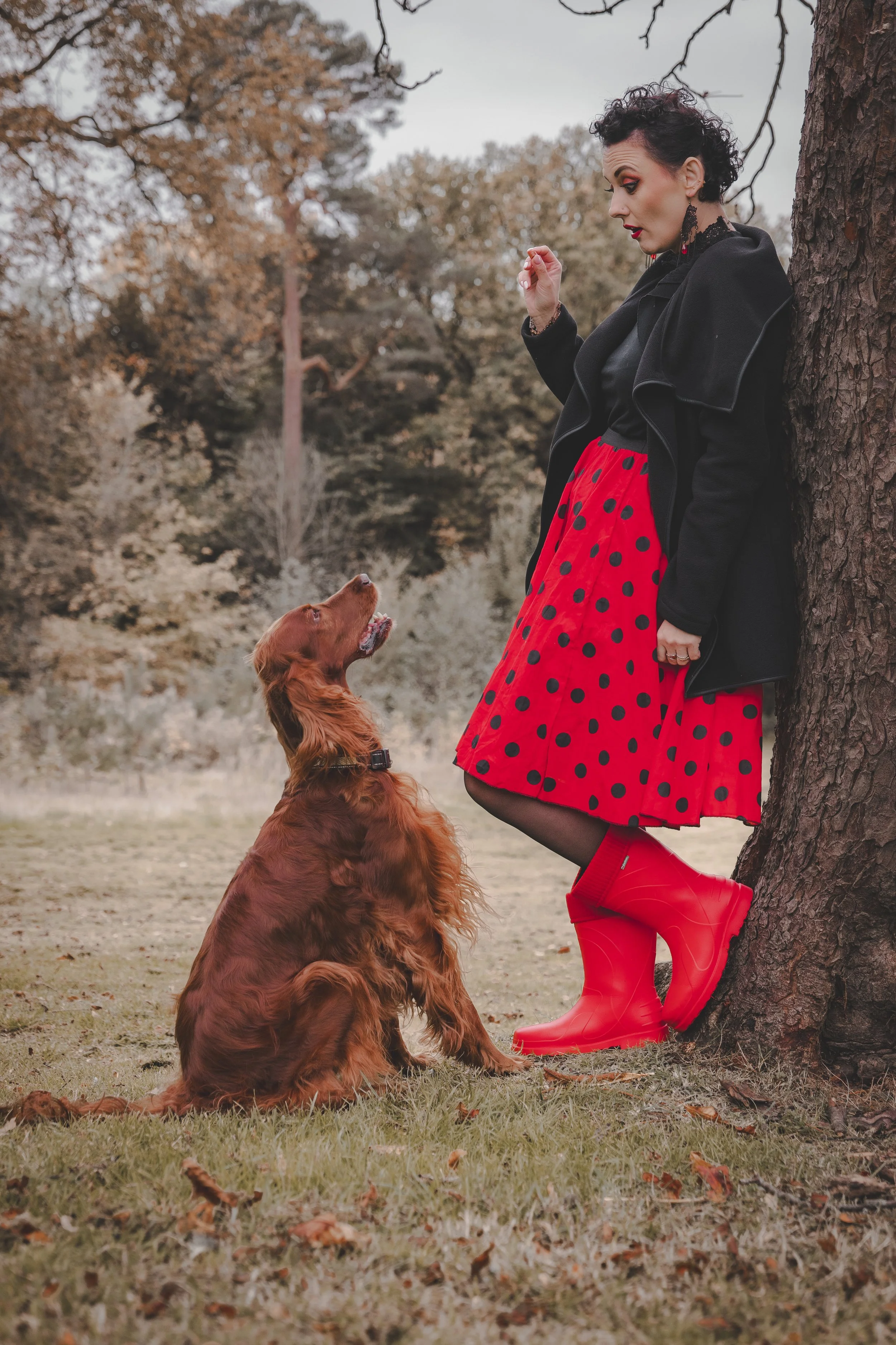 A woman with short dark curly hair wearing a black coat, black dress, red polka dot skirt, and red rubber boots stands leaning against a tree. She is looking down at a sitting golden retriever dog that is looking up at her outdoors in a park with aut