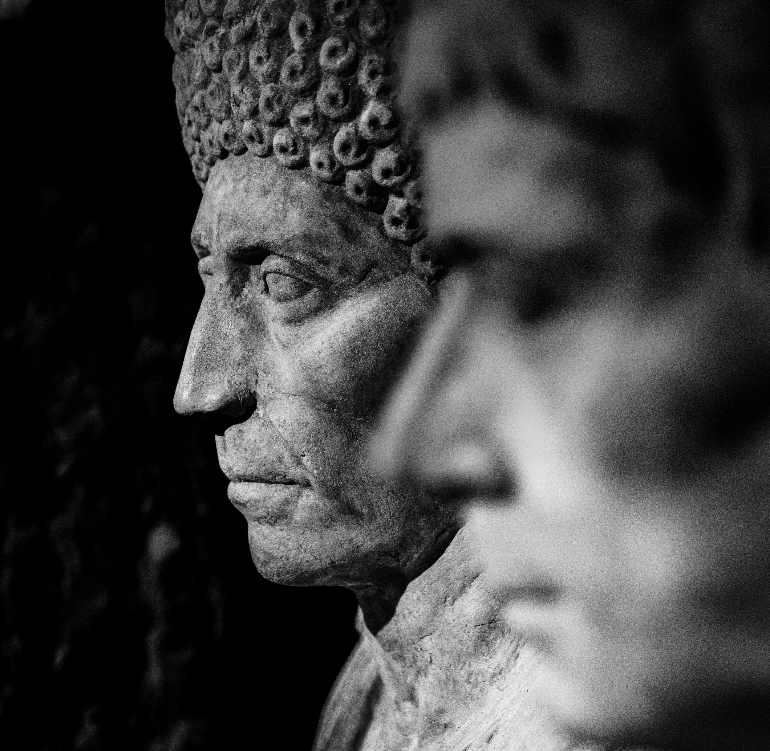 Black and white photograph of two stone sculptures of classical figures, seen in profile with one in the foreground and one in the background.