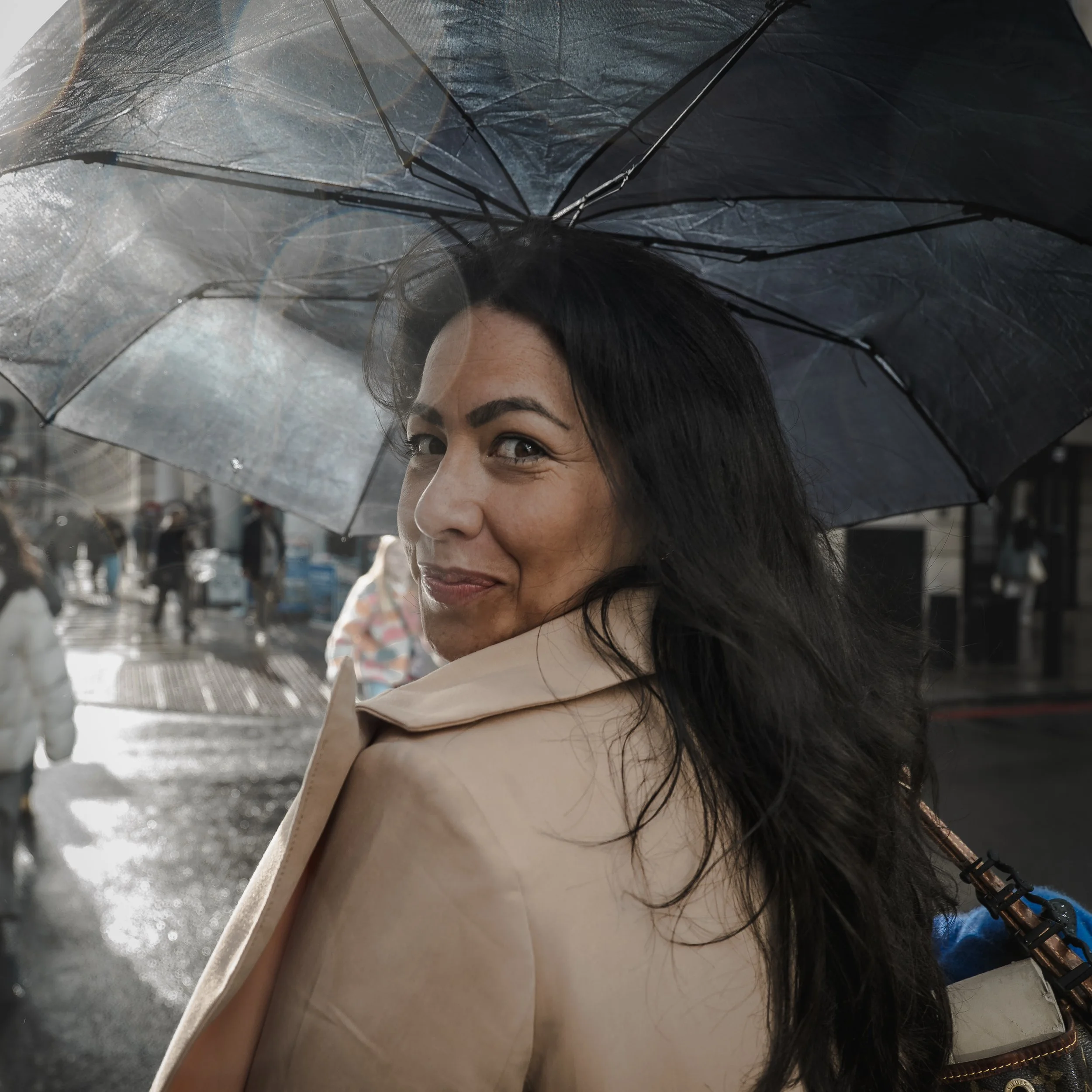 A woman with long black hair smiling and looking over her shoulder on a rainy city street, holding a black umbrella.