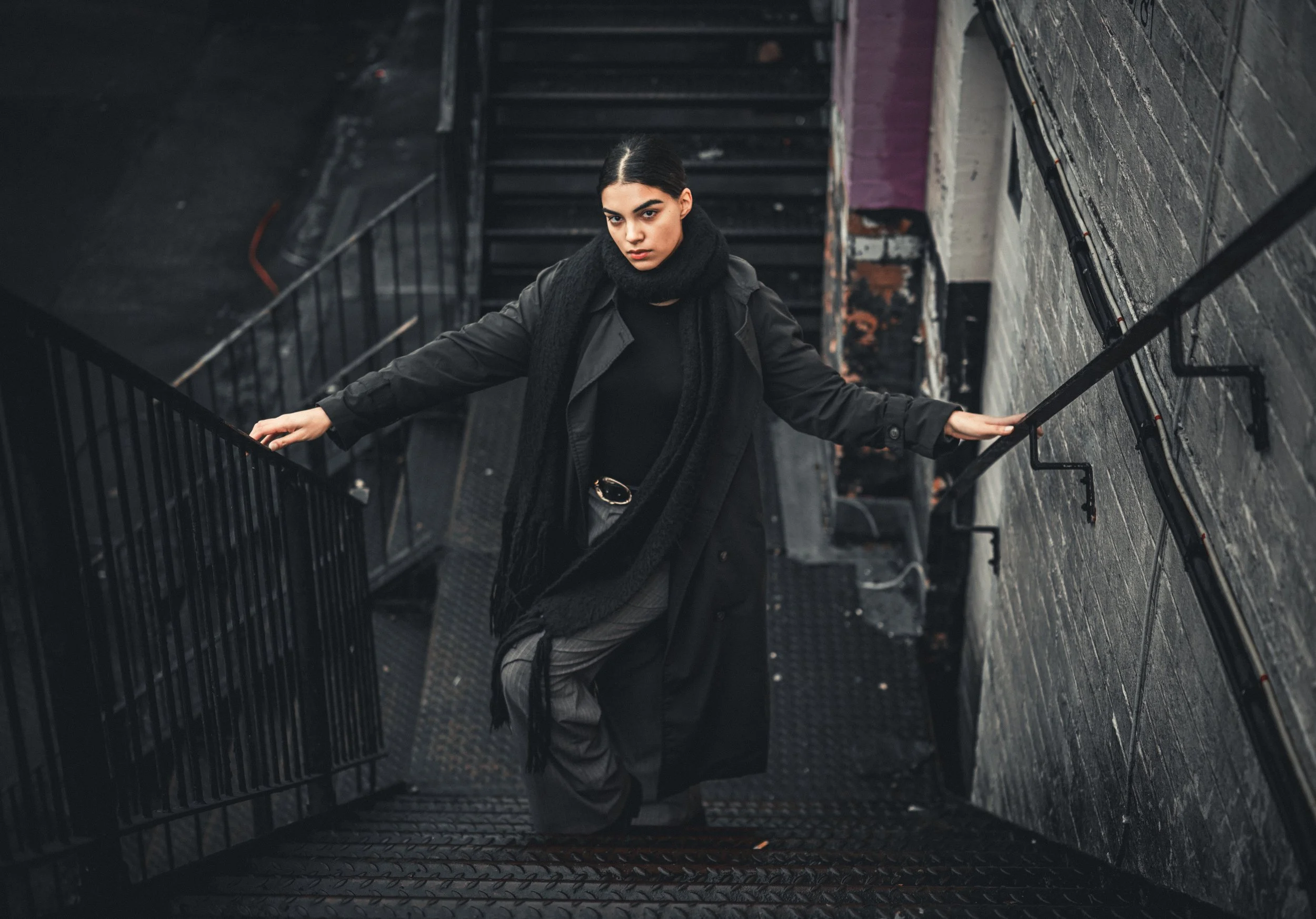 A woman with dark hair, dressed in black, standing on a metal staircase in a dimly lit urban setting, looking up at the camera.