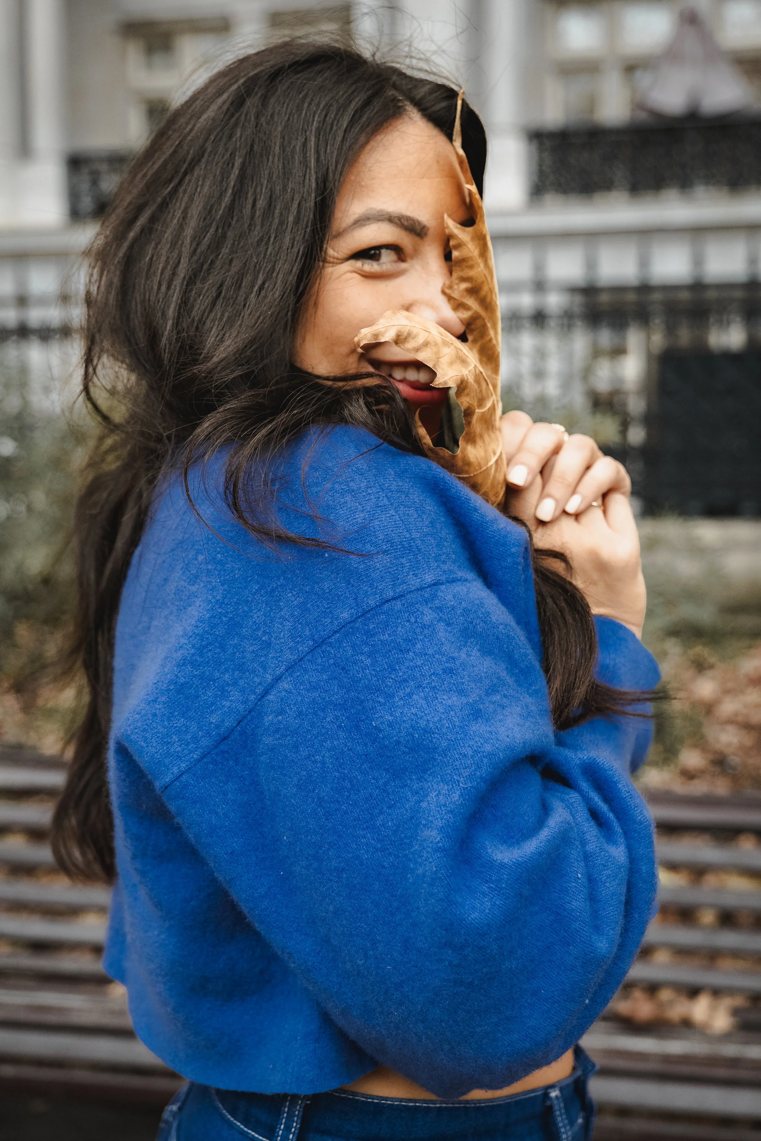 A woman with long dark hair in a blue sweater, holding dried autumn leaves close to her face, smiling with a blurred outdoor background.