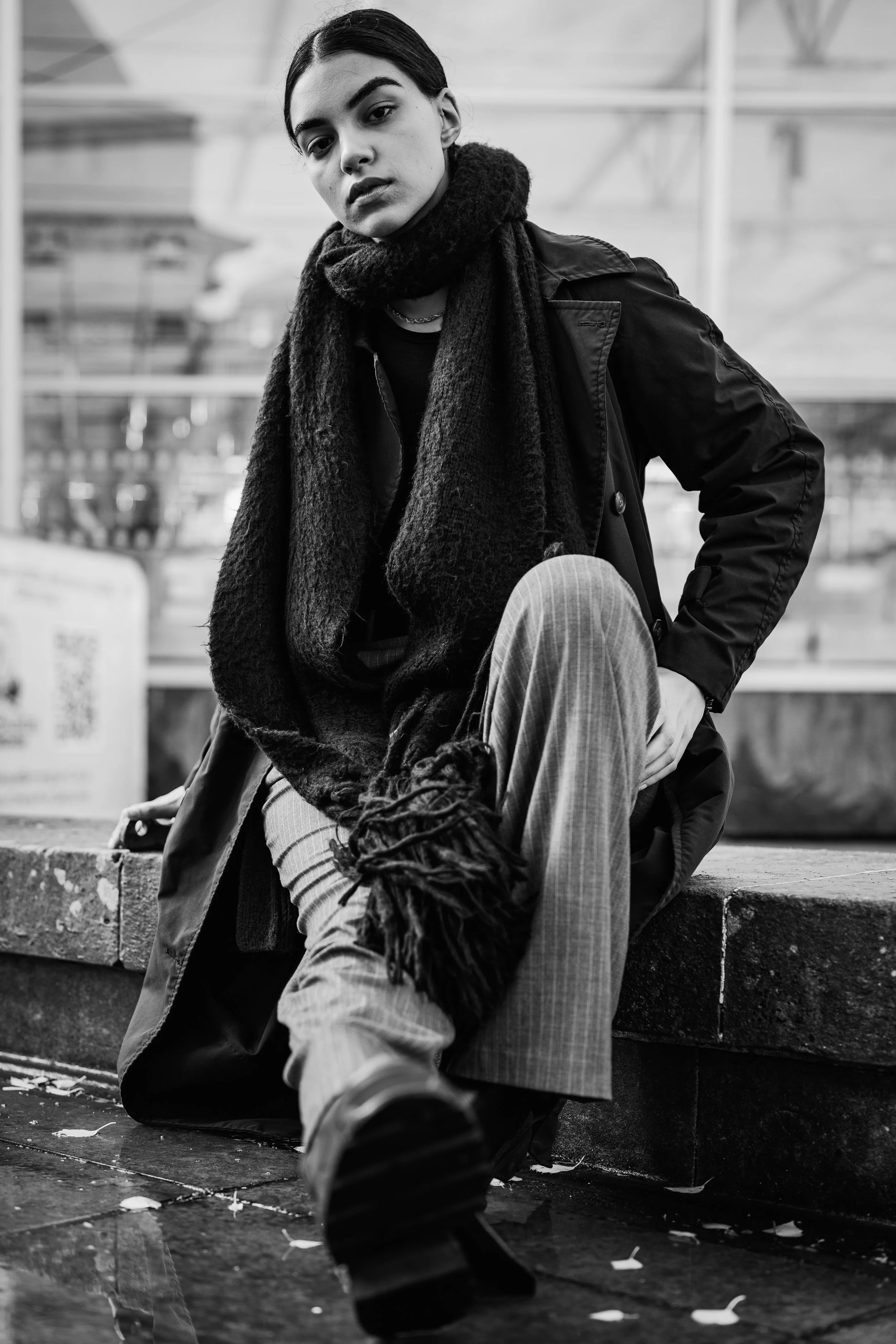 Black and white photo of a woman with dark hair, wearing a scarf, jacket, and patterned pants, sitting on a concrete ledge in an urban environment, looking at the camera.