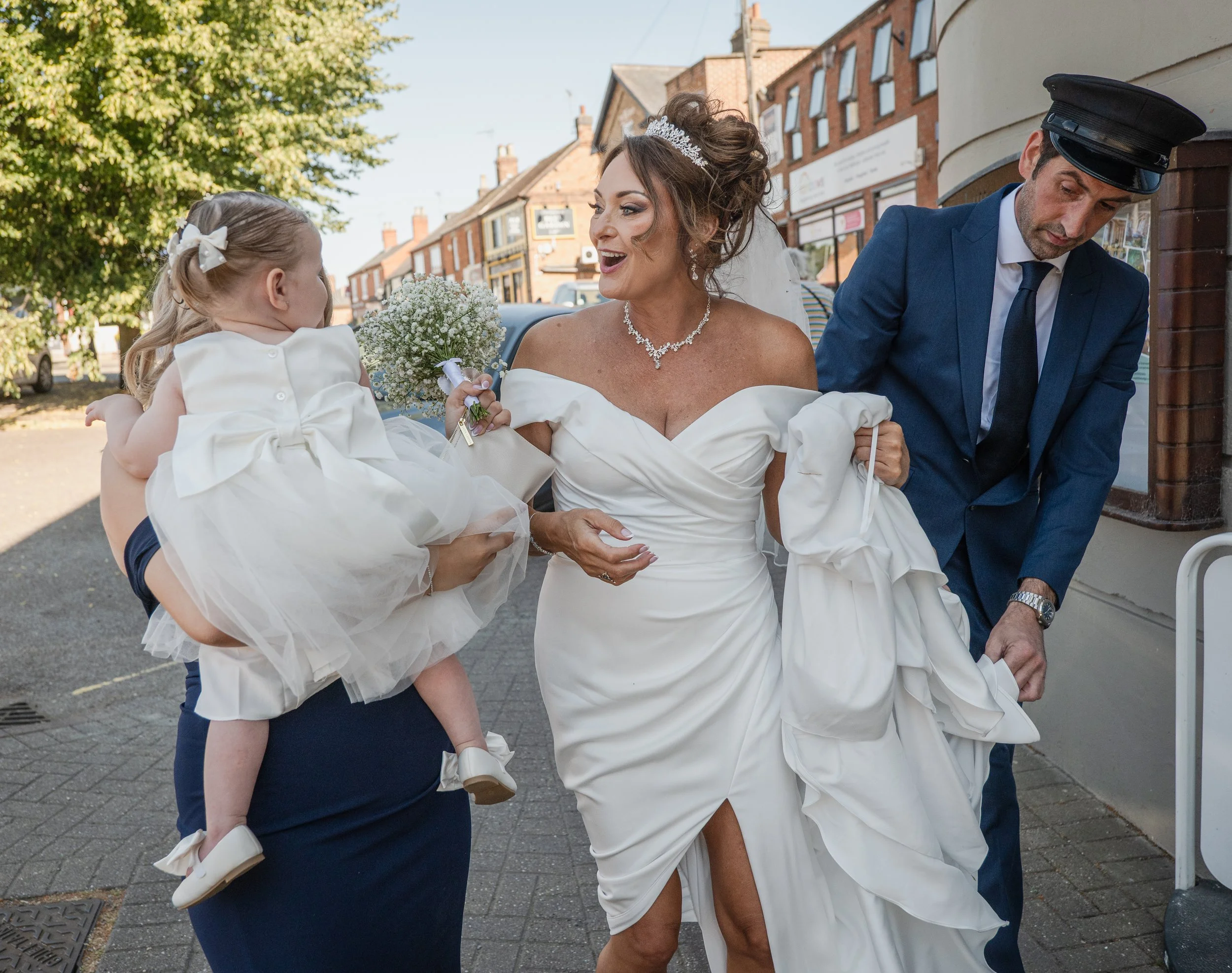 A bride in a white wedding dress and tiara, holding a bouquet, is smiling and talking to a young girl wearing a white dress with a bow, while a man in a blue suit and police cap stands nearby outside on a sunny day.