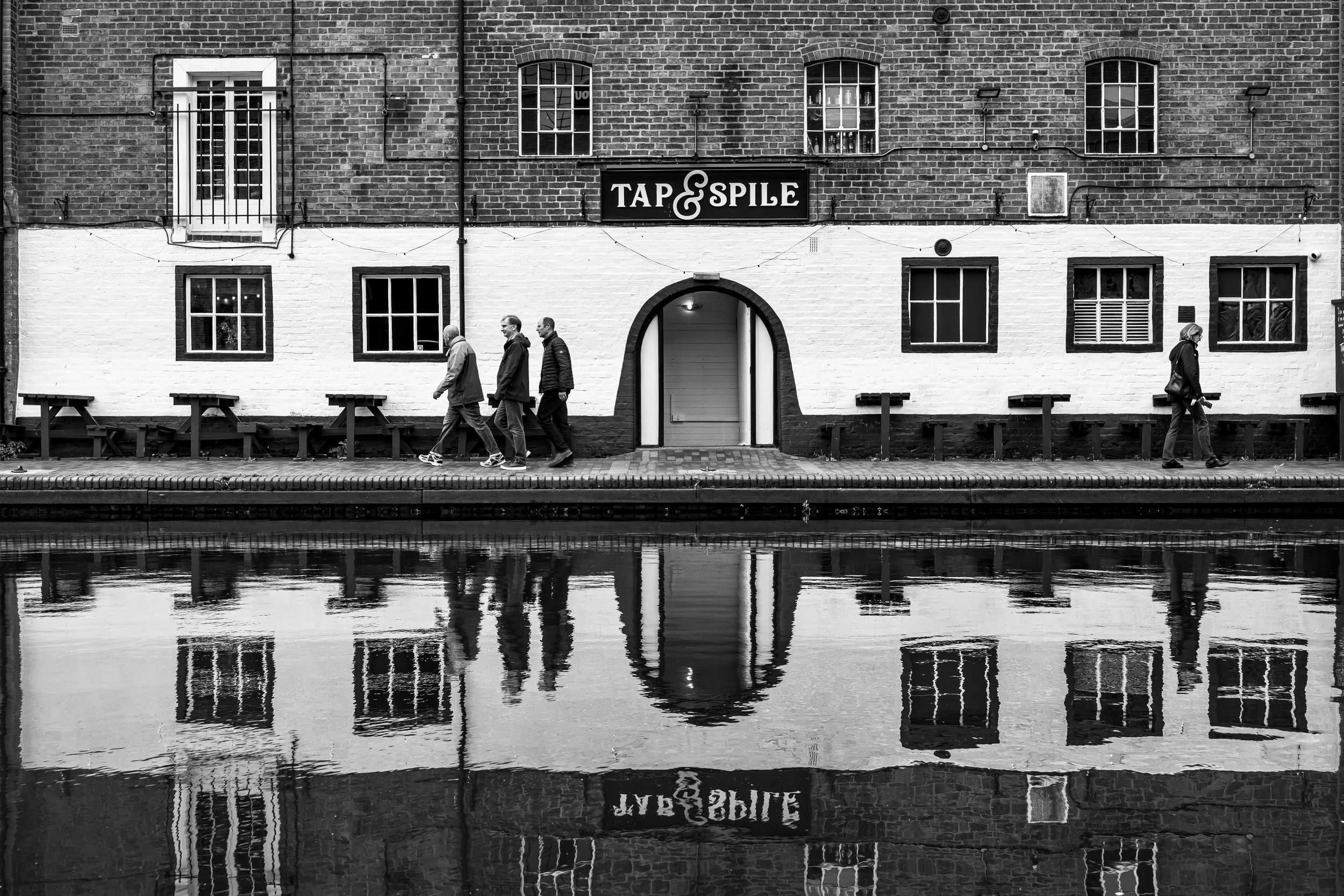 Black and white photo of a riverside bar with a sign reading 'Tap & Spile'. Four people walk on the sidewalk beside the building, reflected in the water in front of them. The building has brick walls, multiple windows, and a central arched door.