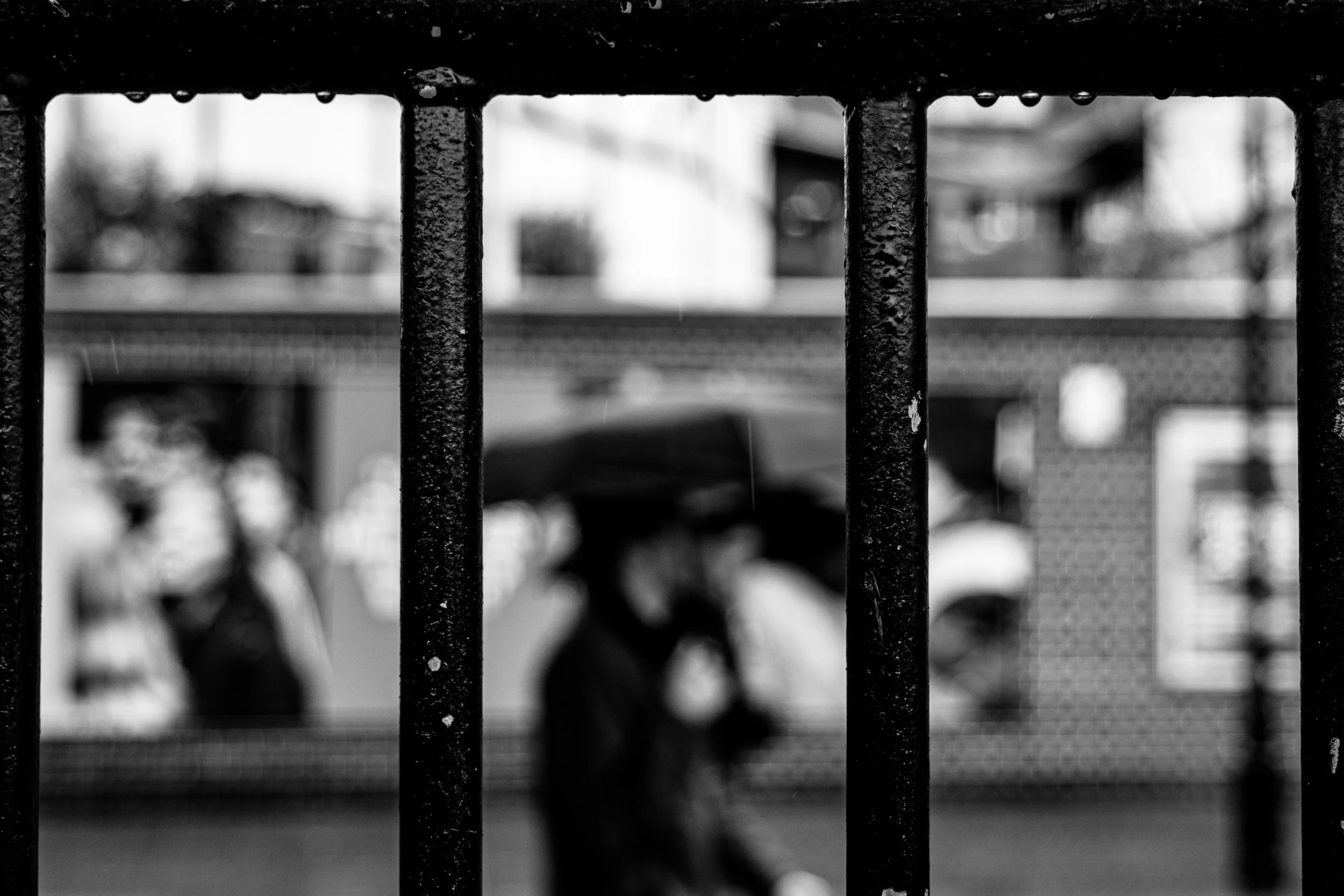 Black and white photograph of a metal fence with vertical bars in focus, with blurred figures of people walking and buildings in the background.