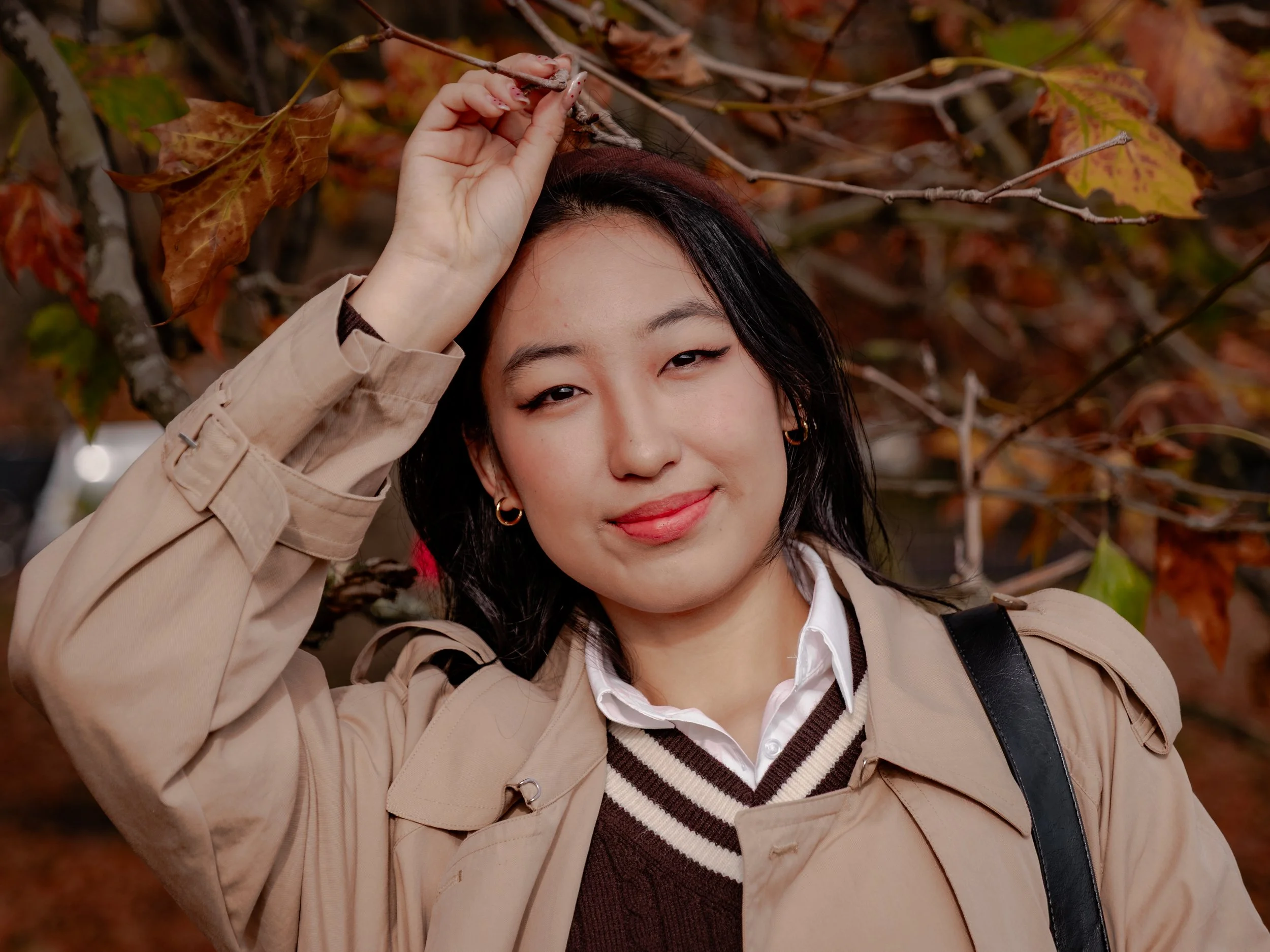 A woman outdoors in autumn, smiling with closed eyes, wearing a beige trench coat and white shirt, standing against a backdrop of fall-colored leaves and branches, with her hand raised to touch a branch.