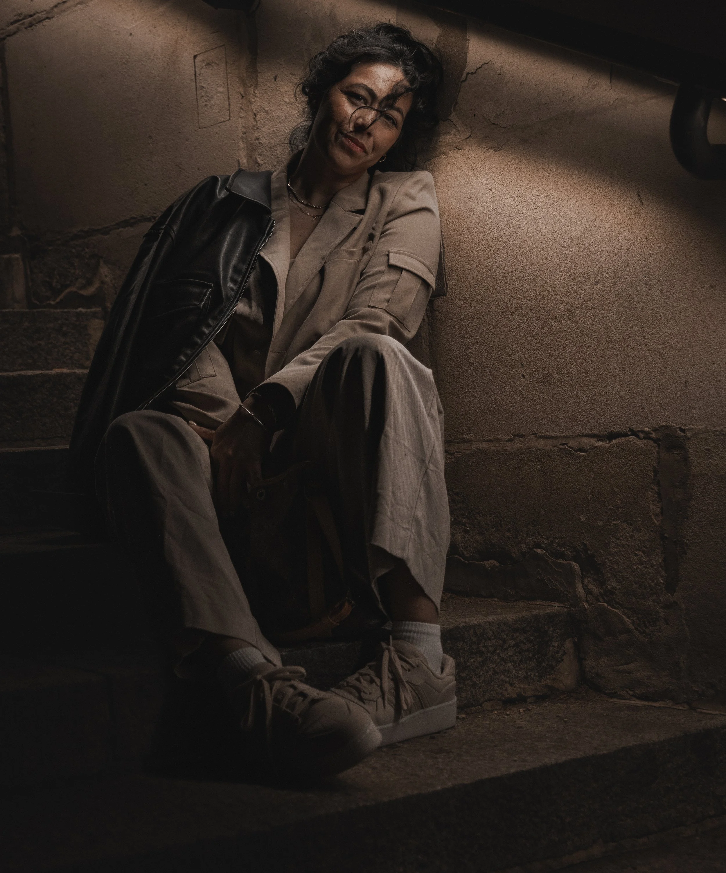 Woman with curly hair sitting on a sidewalk at night, leaning against a stone wall, wearing beige clothing, white sneakers, and a black leather jacket.