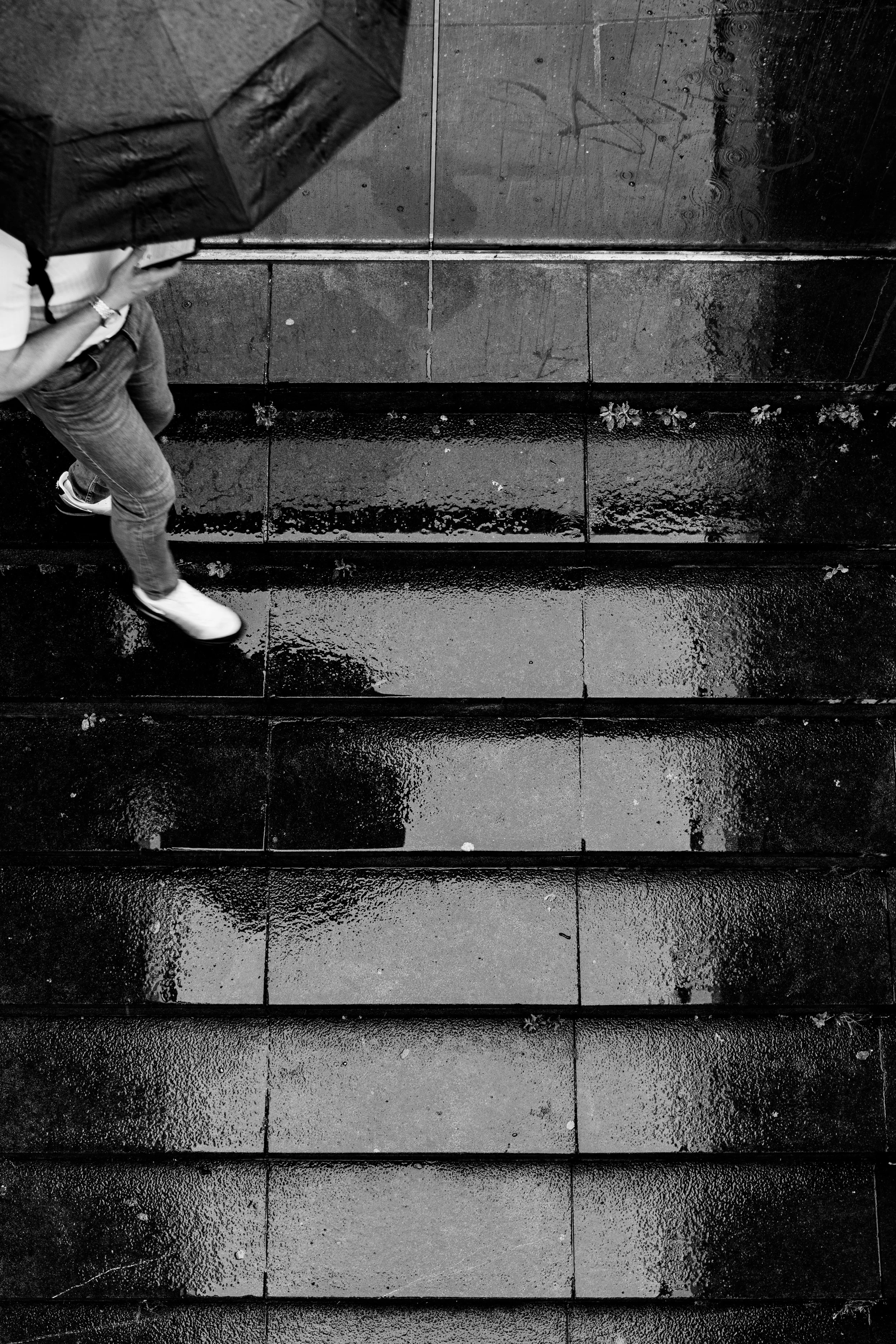 A person holding an umbrella walking on wet pavement with reflections, viewed from above in black and white.