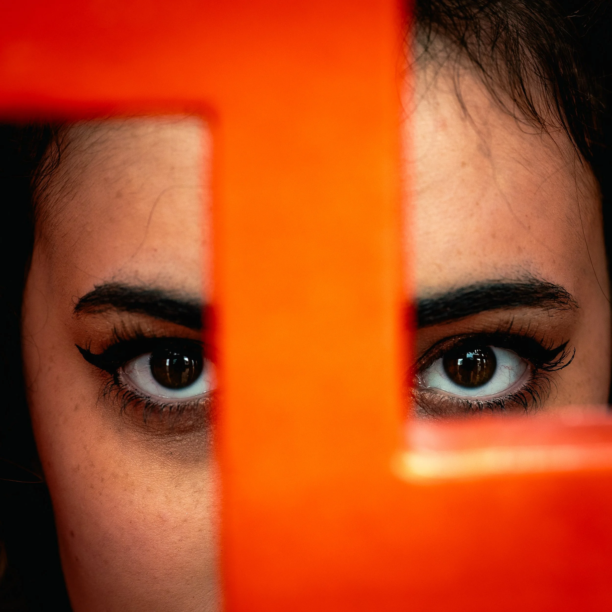 Close-up of a woman's face with brown eyes and makeup, partially obscured by an orange object.