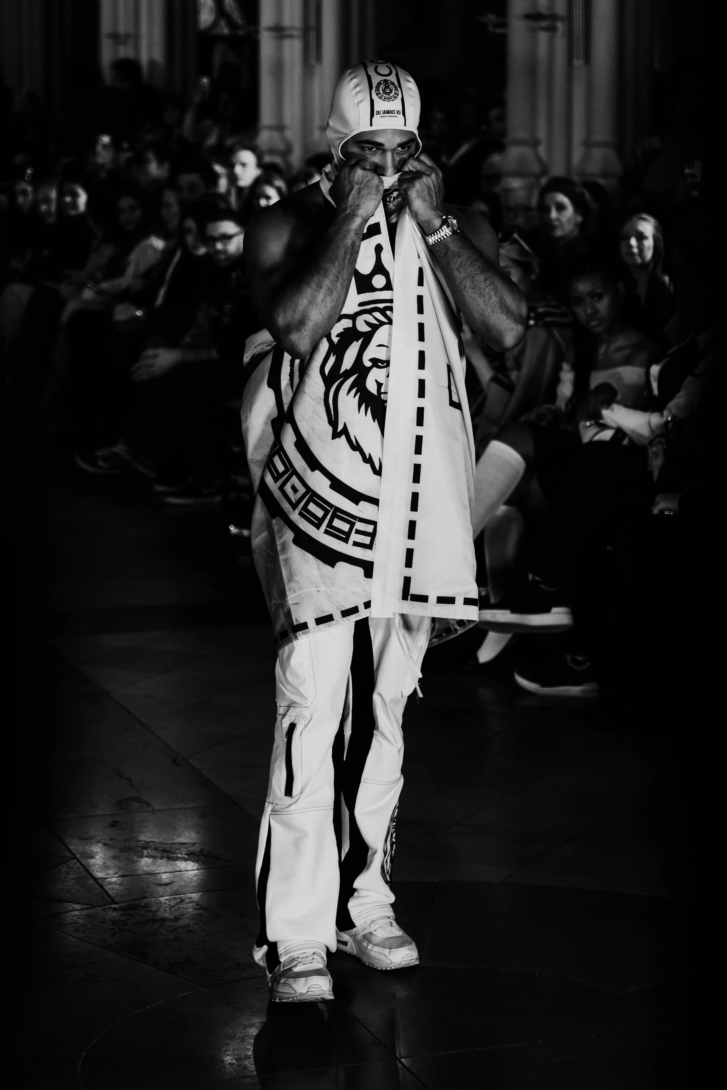 A man at a sporting event, adjusting his shirt or scarf, wearing a hat and athletic clothing, with an audience seated in the background.