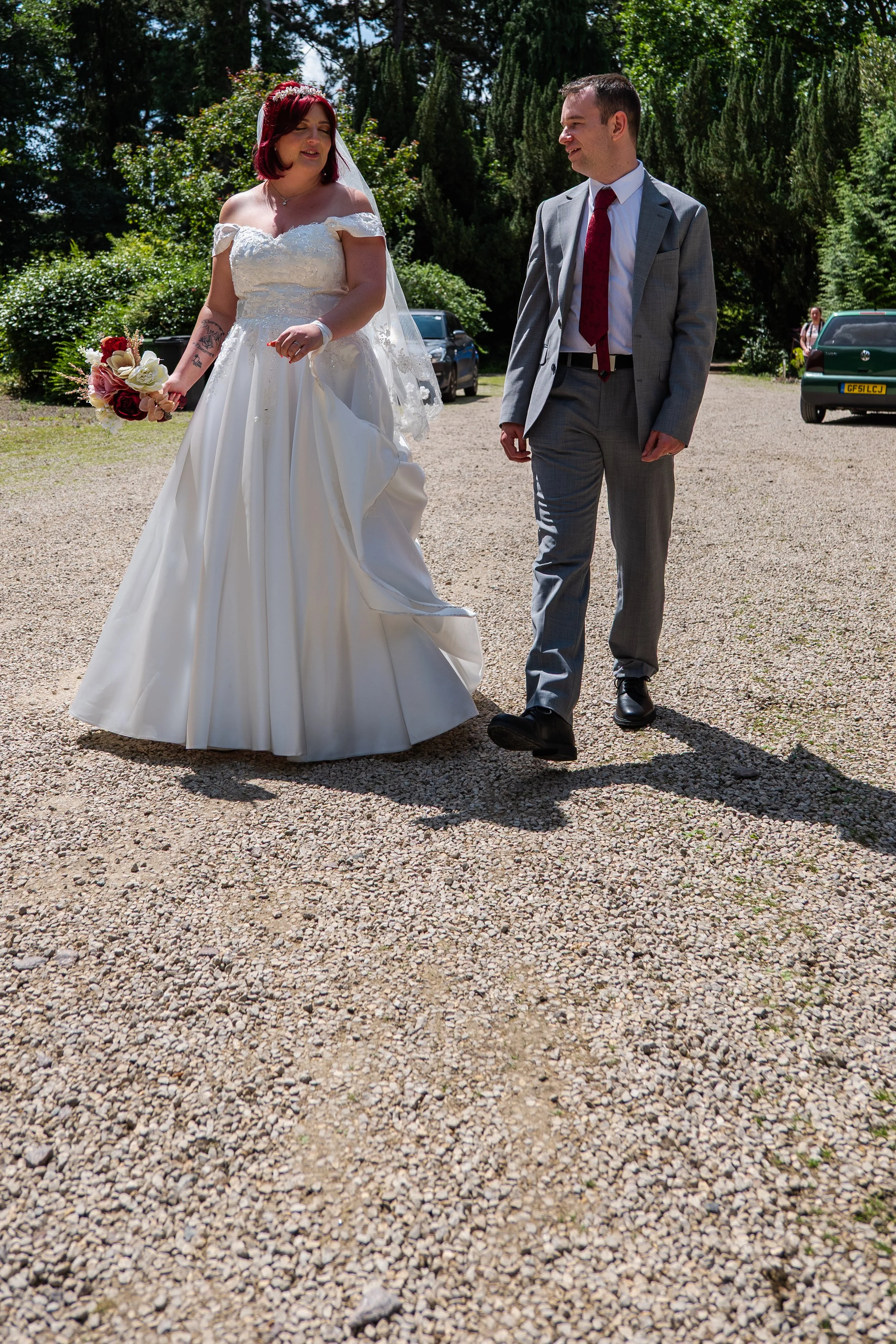 A bride in a white wedding dress and veil holding a bouquet of flowers, and a groom in a gray suit with a red tie, walking outside on a sunny day surrounded by trees and parked cars.