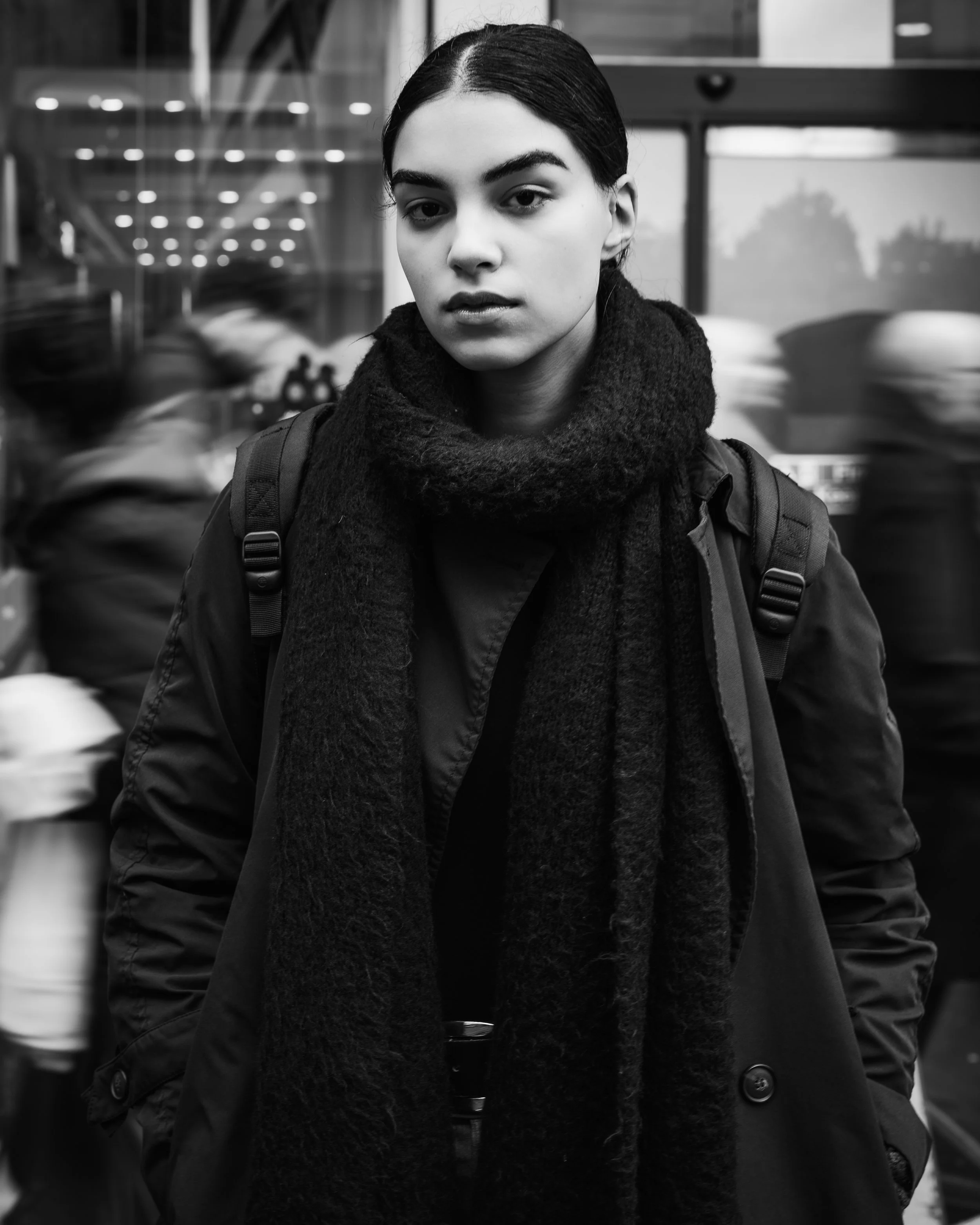 A young woman with dark hair, wearing a dark coat and a large scarf, stands in a busy urban setting with blurred pedestrians in motion behind her.