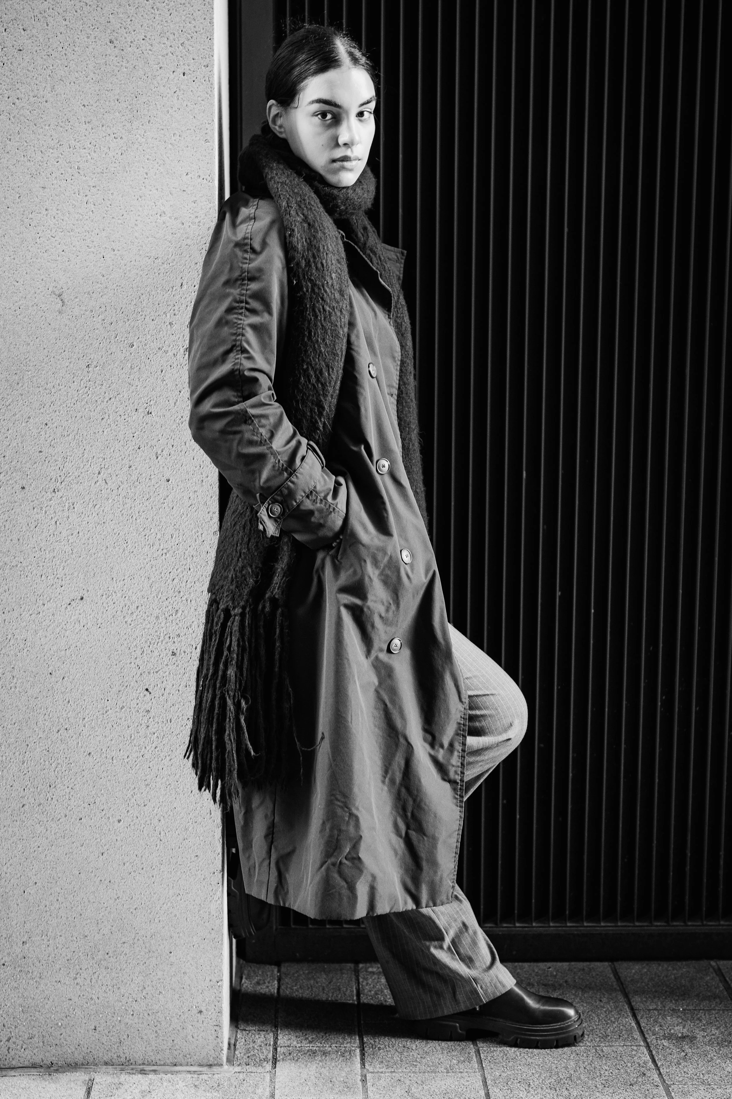Black and white photo of a young woman leaning against a wall, wearing a long trench coat, scarf, pants, and boots, looking directly at the camera.