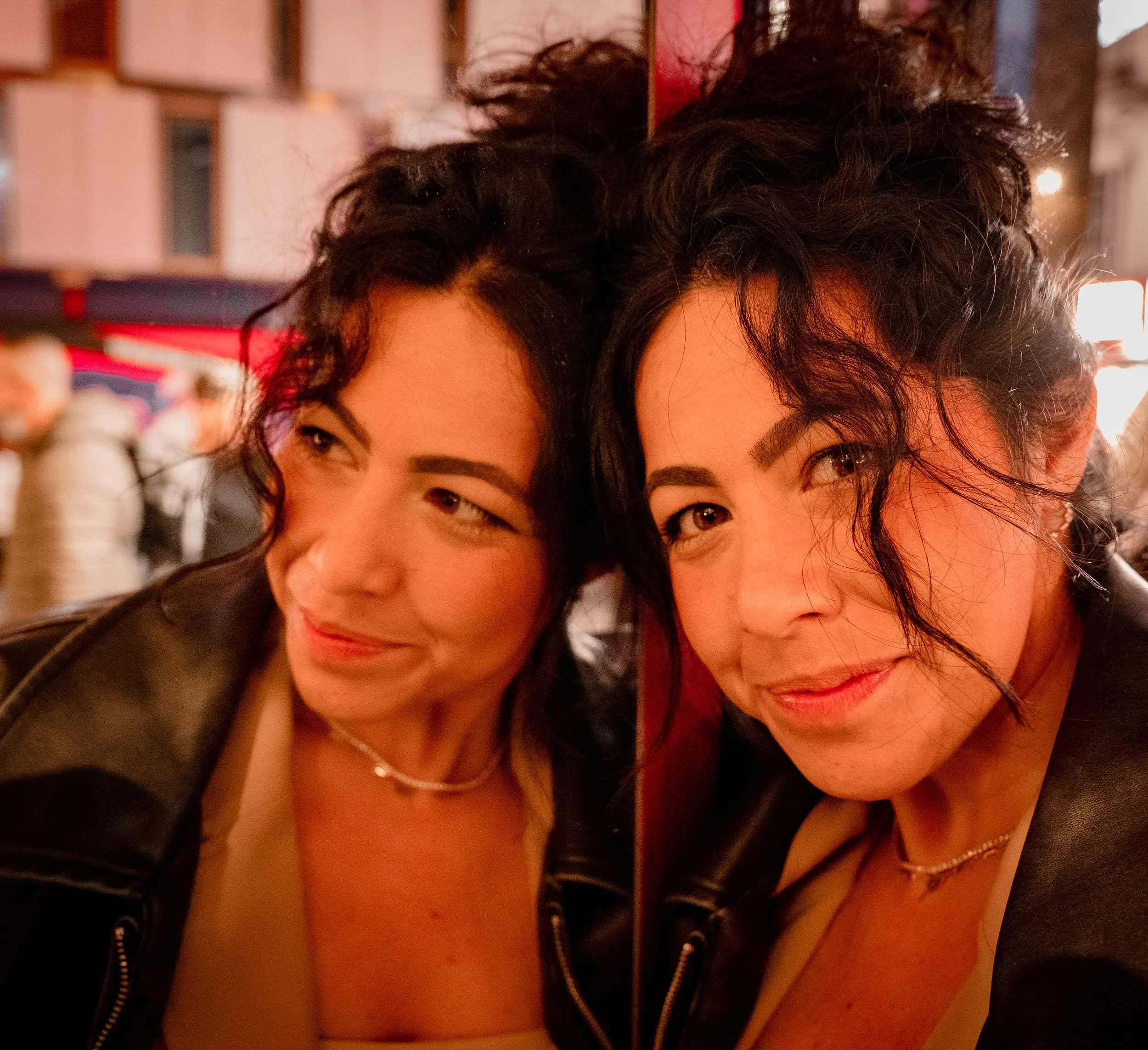 Two women with dark, curly hair taking a selfie at night, smiling, in an urban setting with blurred people in the background.