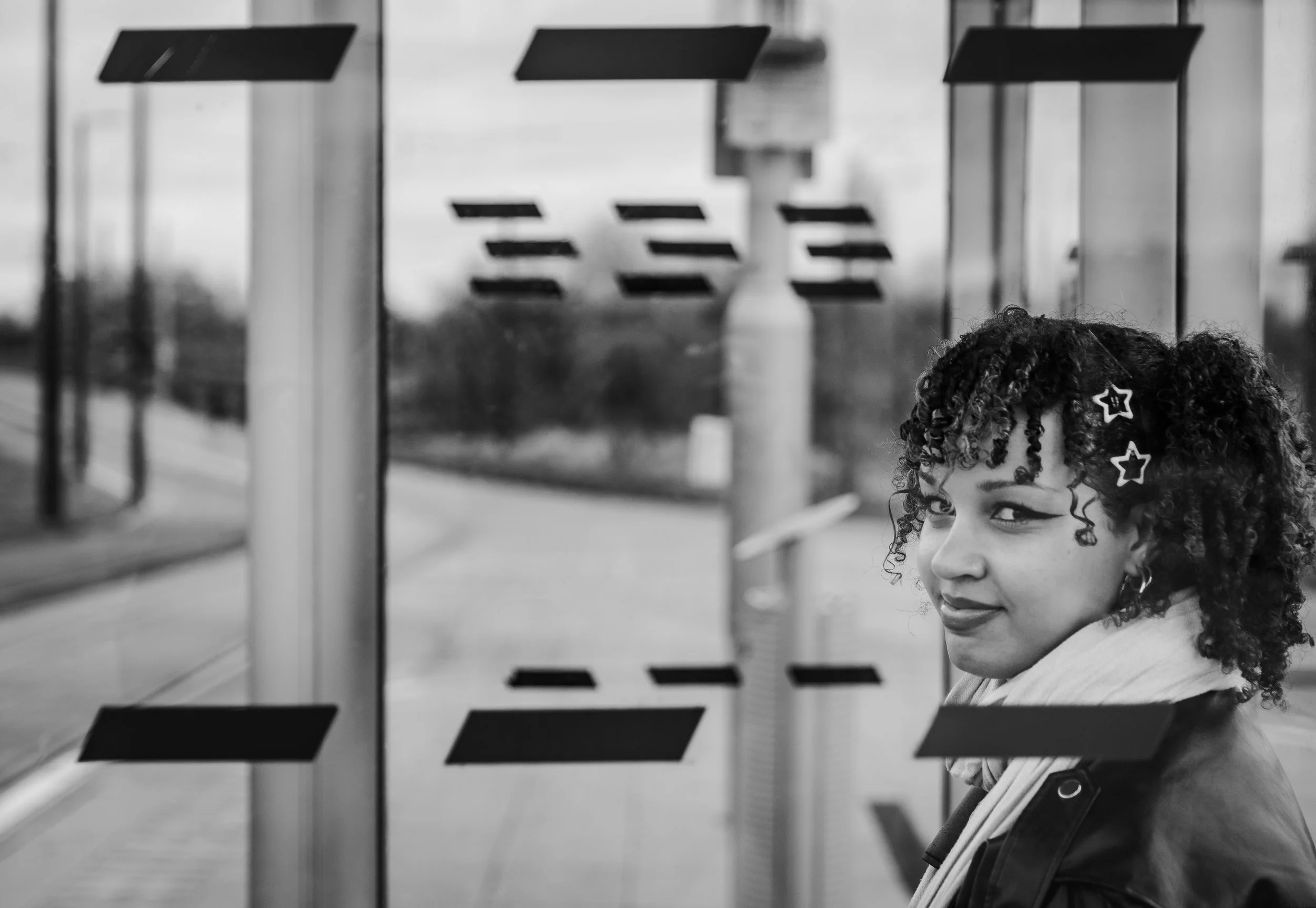 A young woman with curly hair and star-shaped hair clips standing at a bus stop, looking over her shoulder, with a city street visible through the glass behind her.