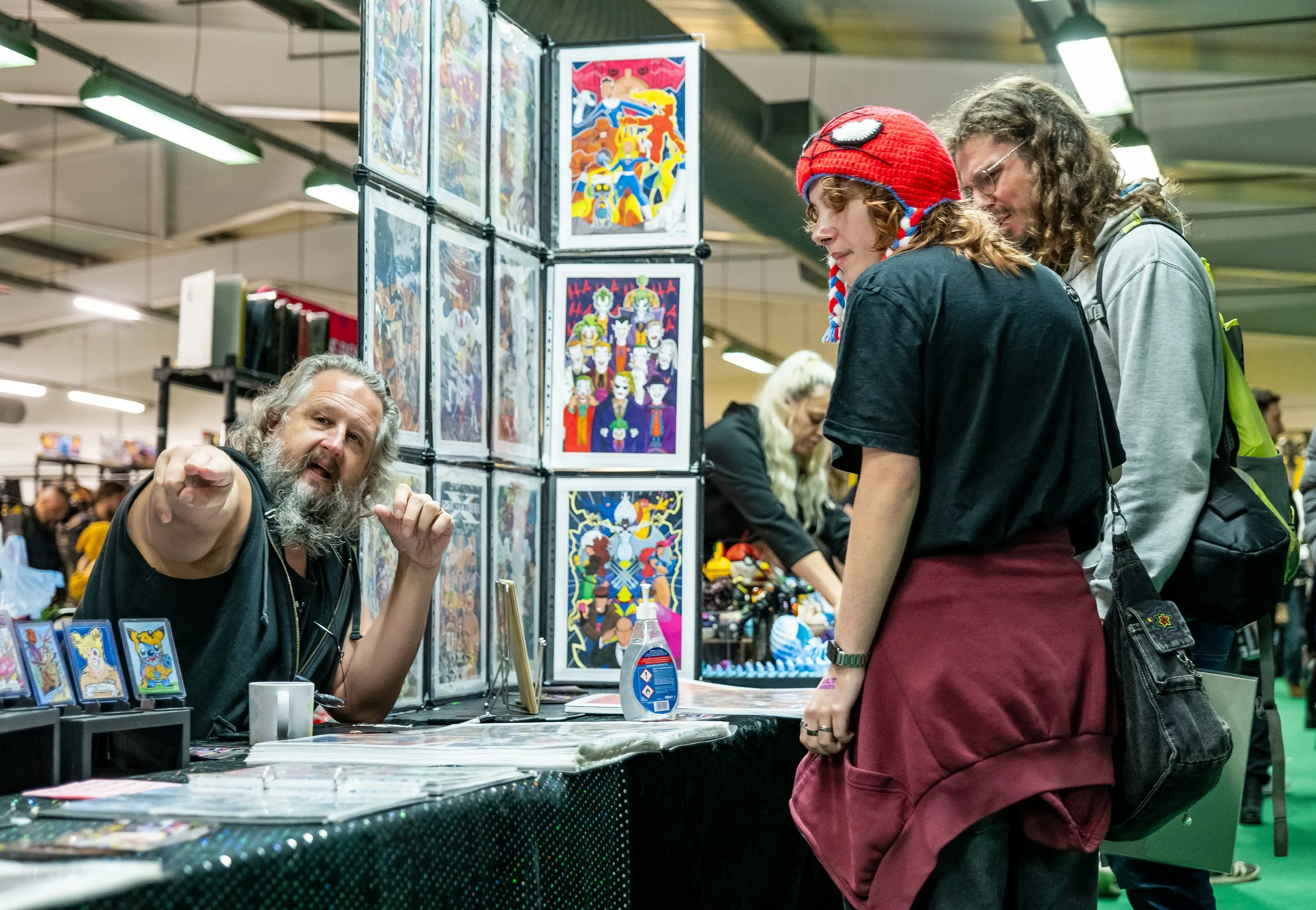 A man with long gray hair and a beard is sitting at a table with comic art prints, talking to two young women who are looking at the artwork. The setting appears to be a comic convention or art fair.