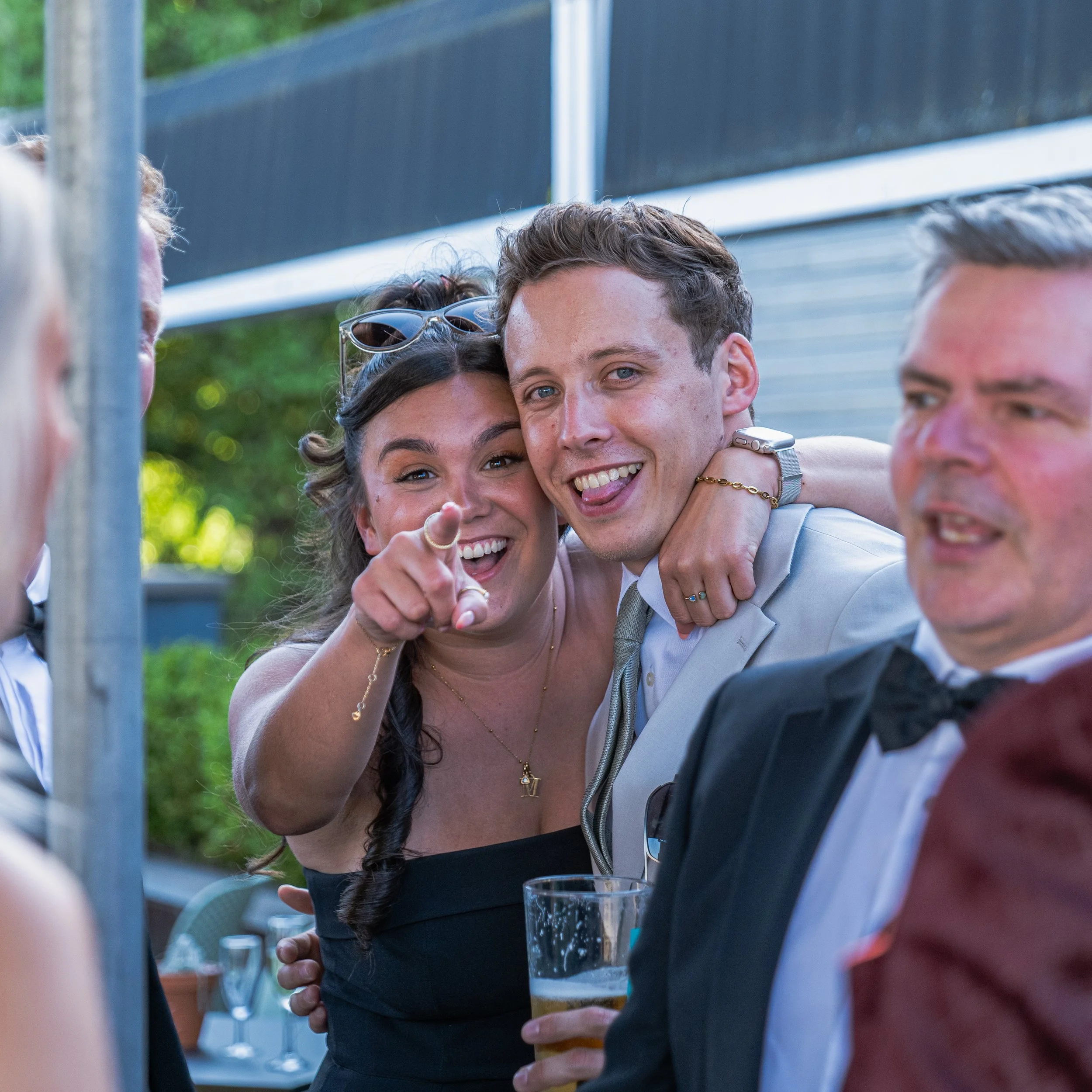 A group of friends at a formal event outdoors, smiling and enjoying drinks, with some dressing in tuxedos and women in dresses.
