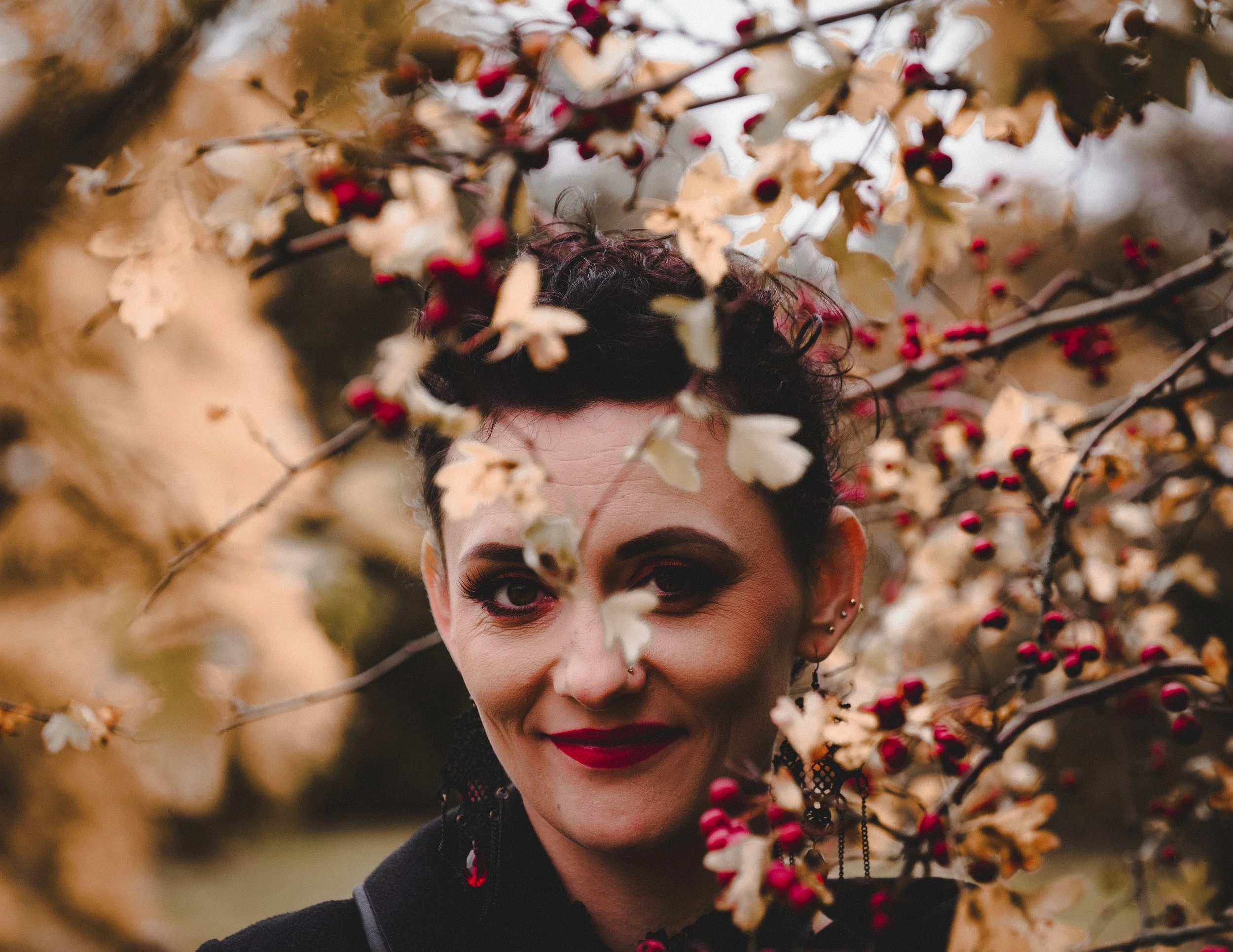 A woman with dark makeup, short dark hair, and red lipstick, peeking through branches with dry beige leaves and small red berries, smiling at the camera.