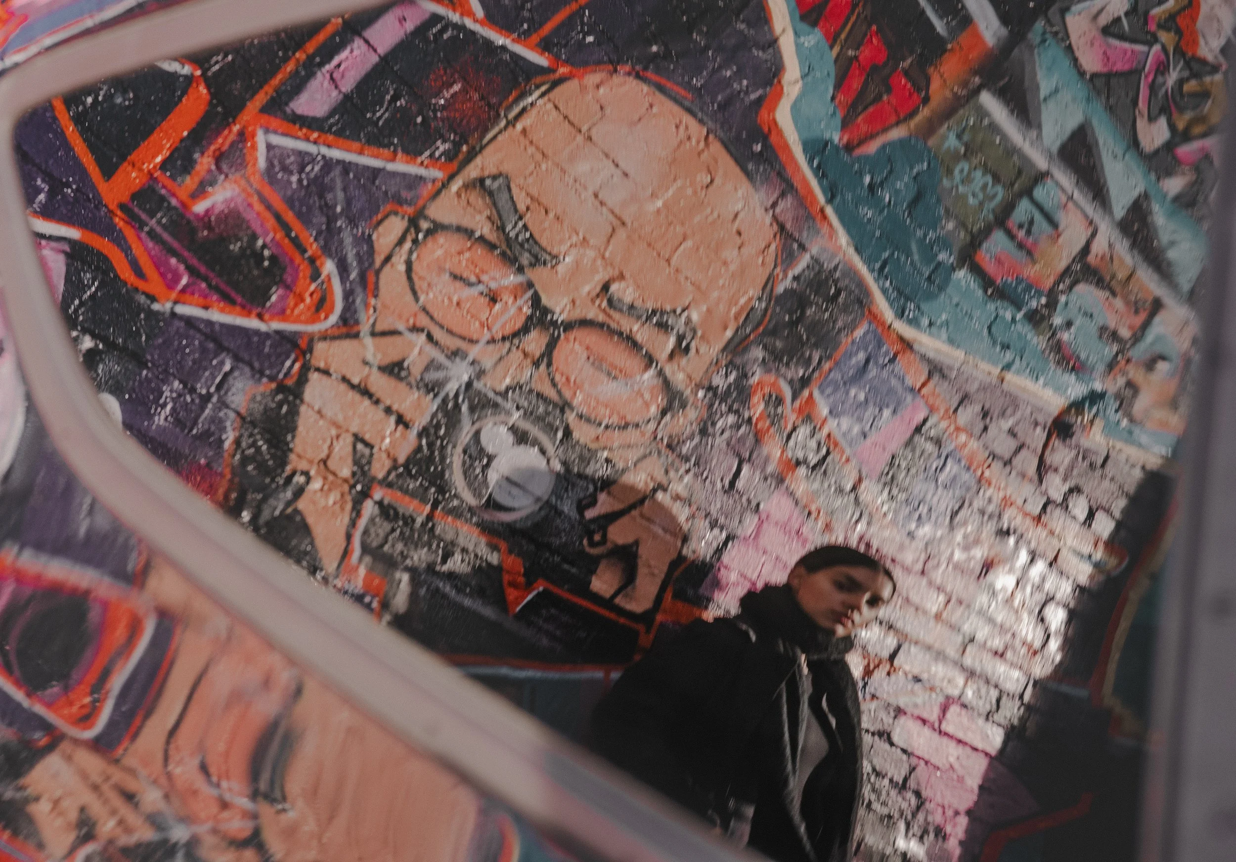 A woman with dark hair pulled back, wearing a black jacket, stands in front of a vibrant, colorful mural on a brick wall. The mural features abstract shapes, graffiti, and a portrait of a man with glasses.