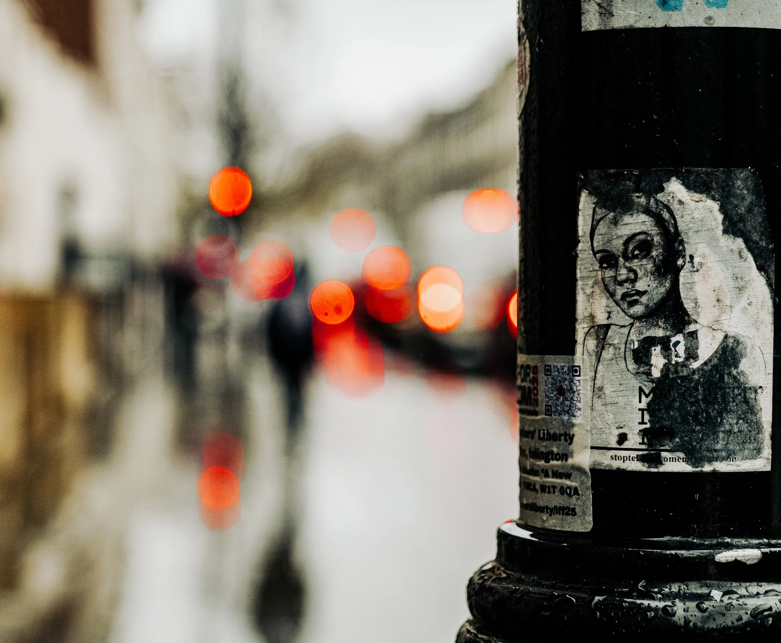 A black pole with a weathered black-and-white sticker of a woman with short hair and a serious expression, on a rainy city street with blurred pedestrians and red bokeh lights in the background.
