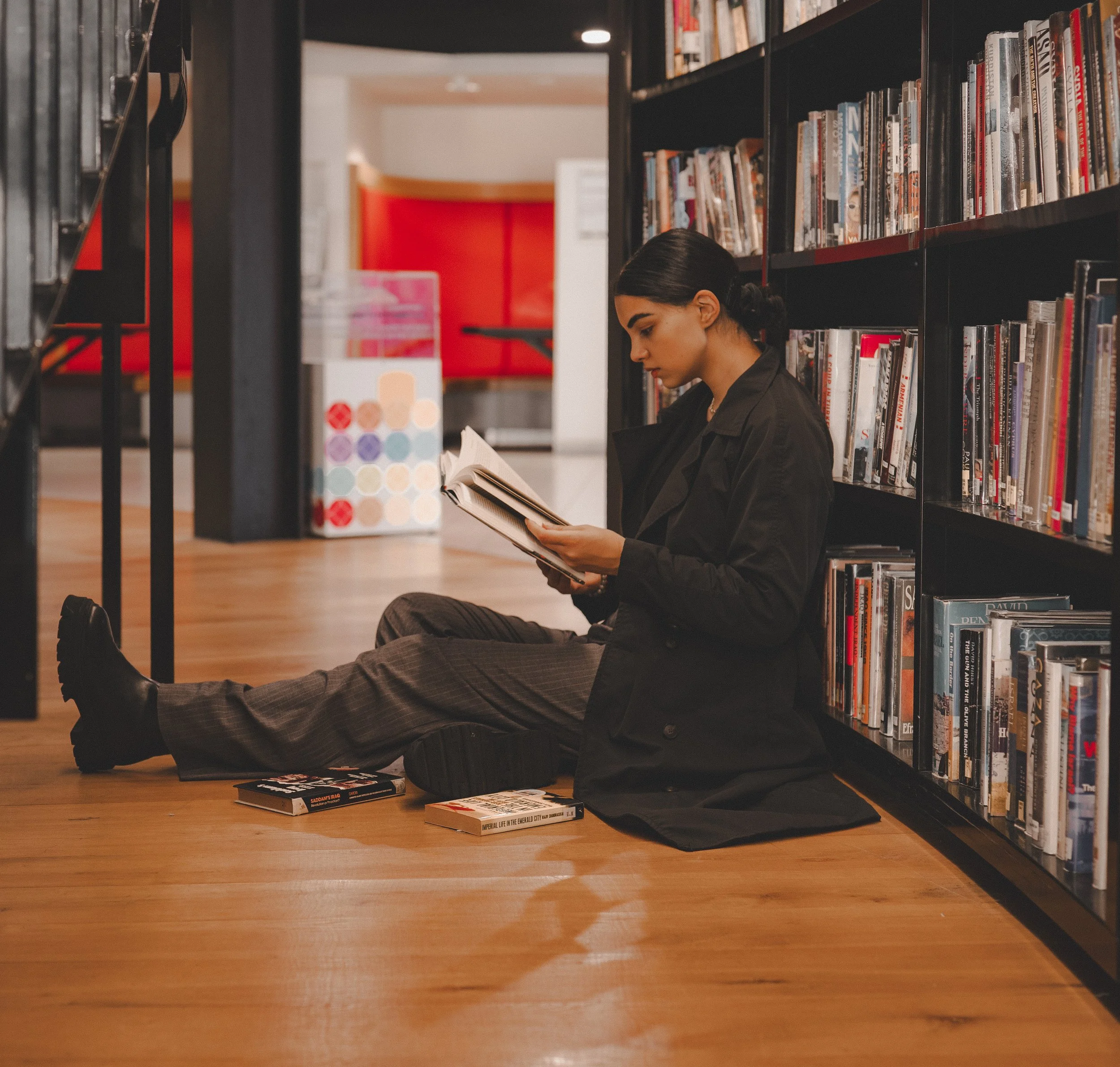 A woman sits on a wooden floor leaning against a bookshelf in a library, reading a book. She is dressed in dark clothing, and her hair is tied back. Several books are scattered on the floor around her.