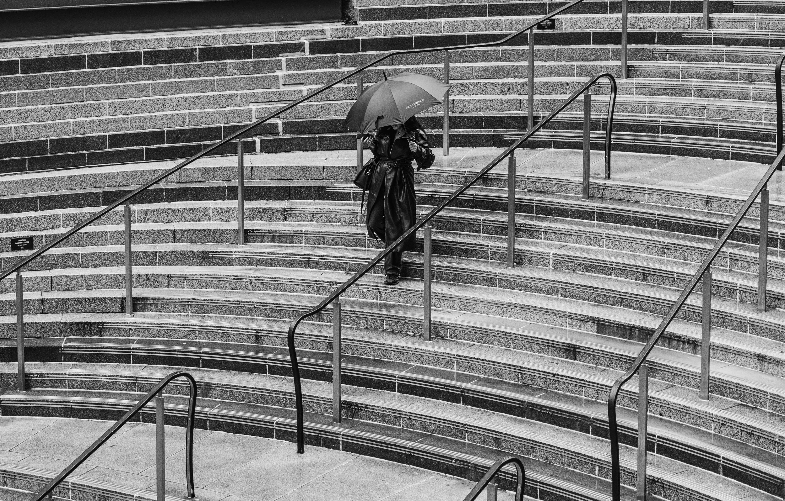 A person walking up outdoor stone stairs with a black umbrella, wearing a dark coat and pants, with metal handrails on either side, in a rainy setting.