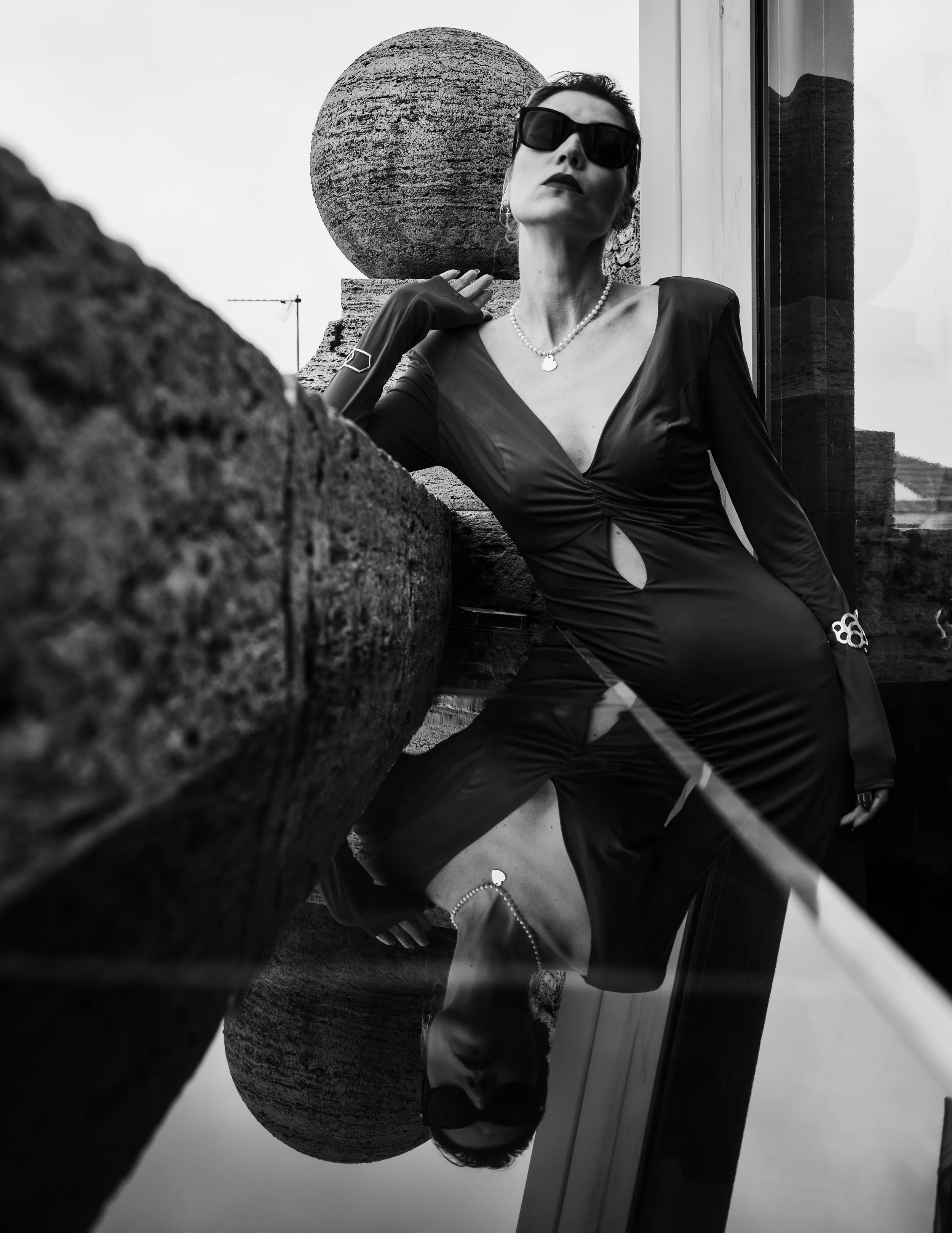 A woman wearing sunglasses, a black dress with a keyhole cutout, and jewelry, leaning on a stone ledge outside next to a glass window. The photo is black and white and is reflected in a puddle below.