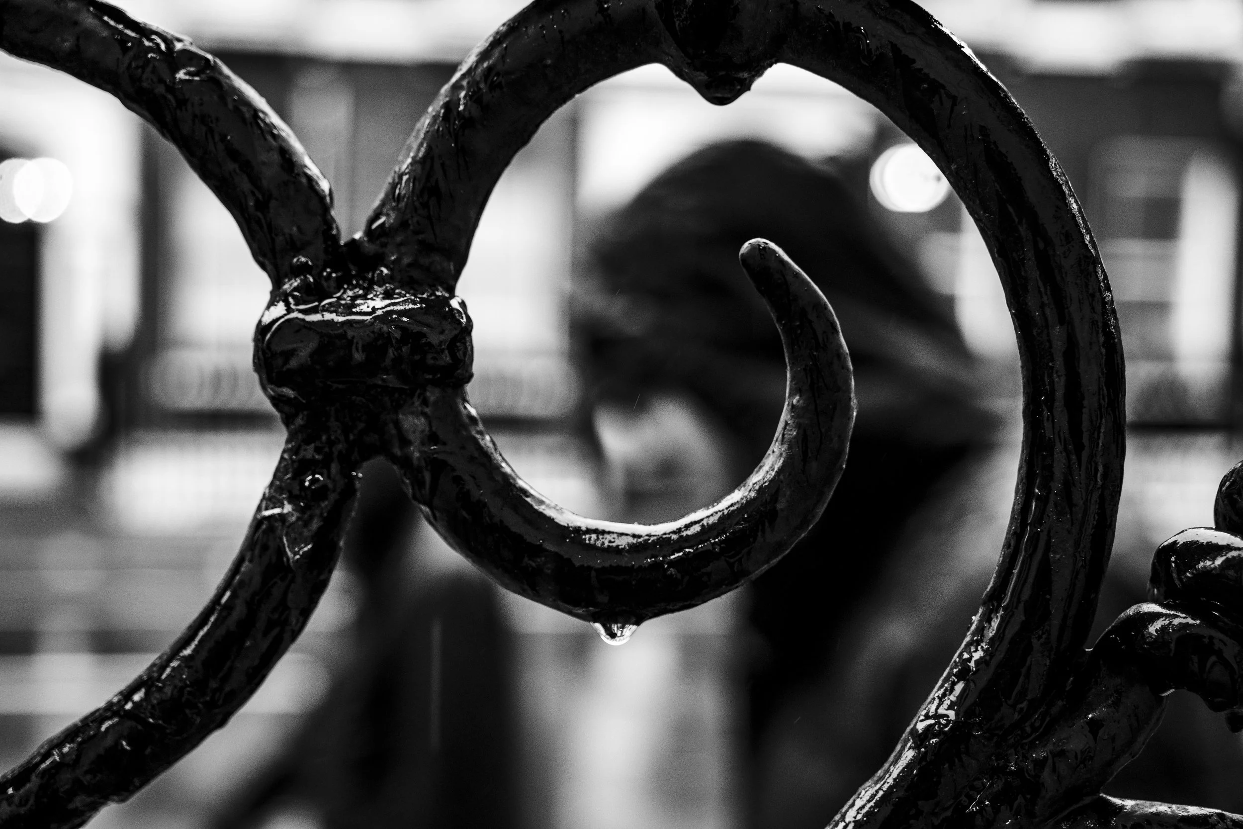 Black and white close-up of a decorative iron railing with a heart-shaped design, and a blurred person in the background.