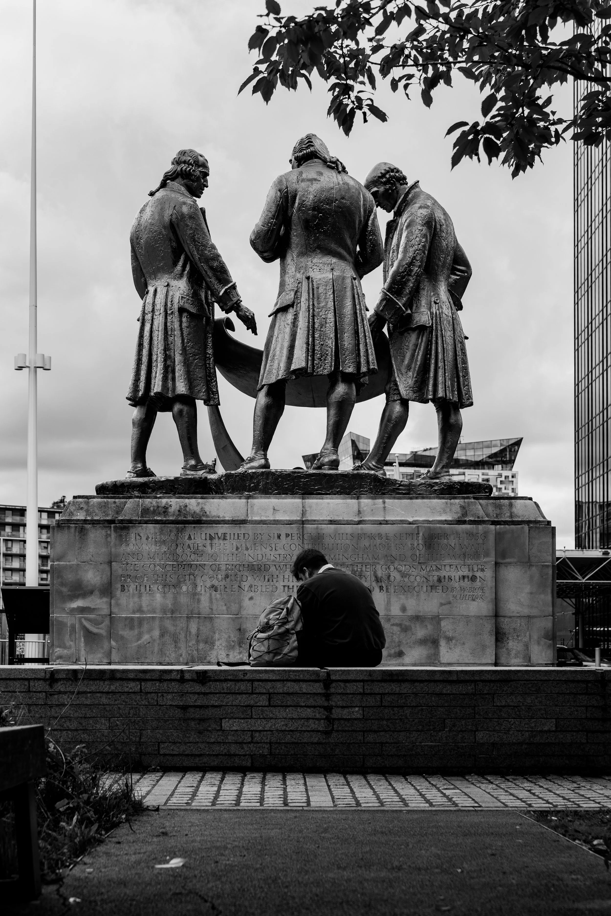A person sitting on a low wall reading a book underneath a large bronze statue of three historical figures, with modern buildings in the background.
