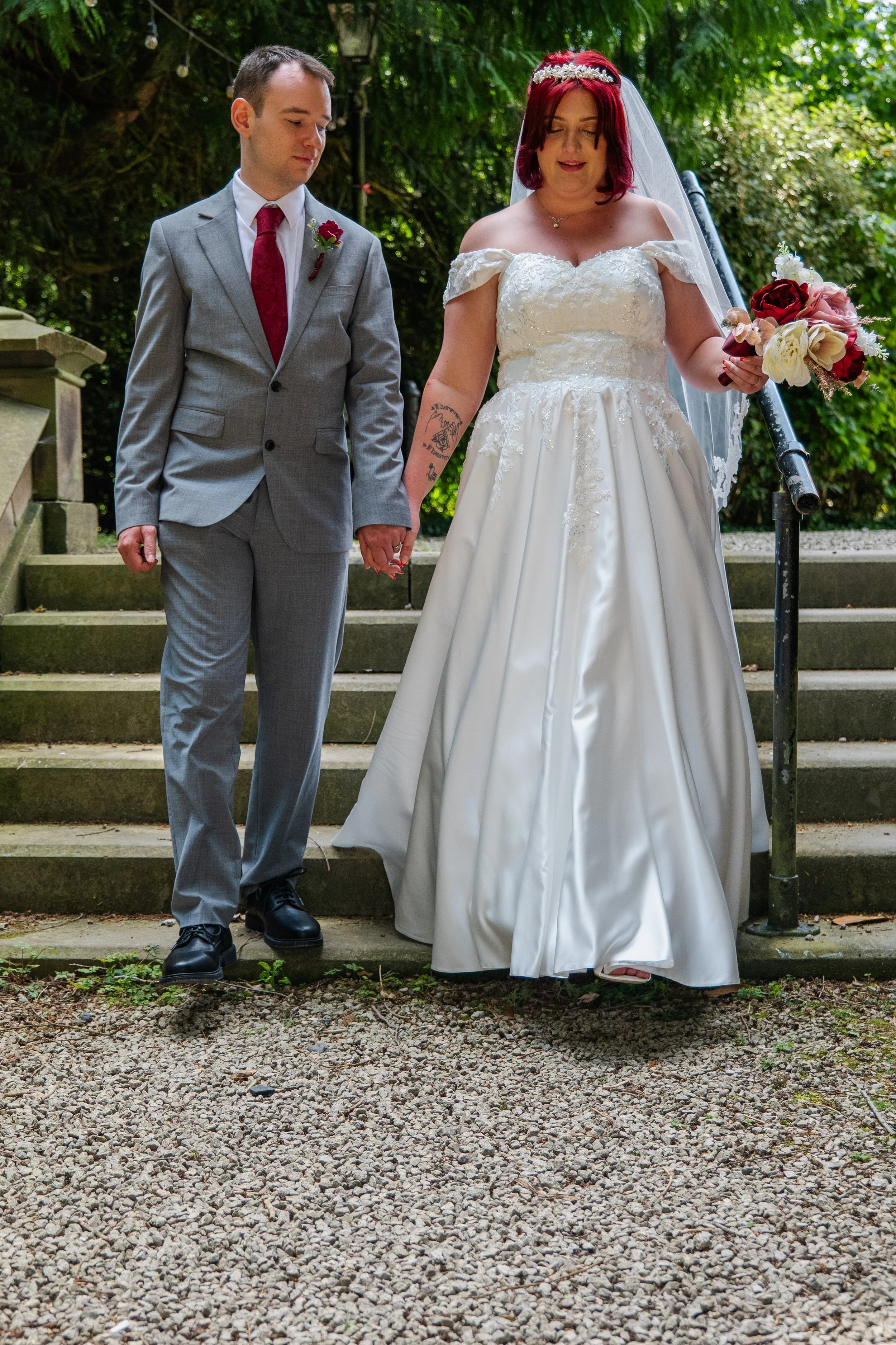 A bride and groom holding hands on outdoor wedding steps, with the bride wearing a white strapless gown and veil, holding a bouquet of roses, and the groom in a gray suit with a red tie and boutonniere, standing under green trees.