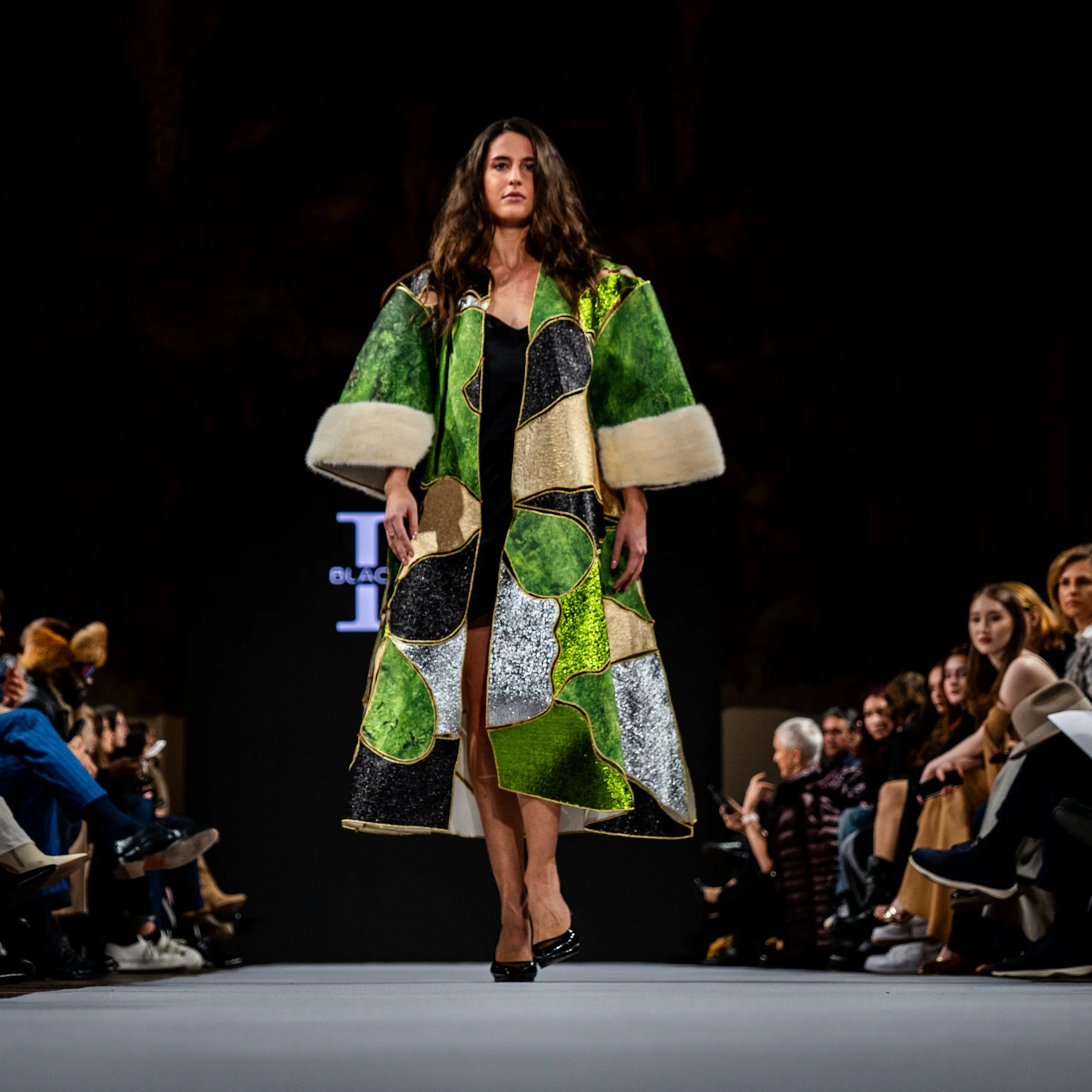 A female model walking on a runway during a fashion show, wearing a long, colorful coat with abstract patchwork in green, black, gold, and silver, and with large white fur cuffs.