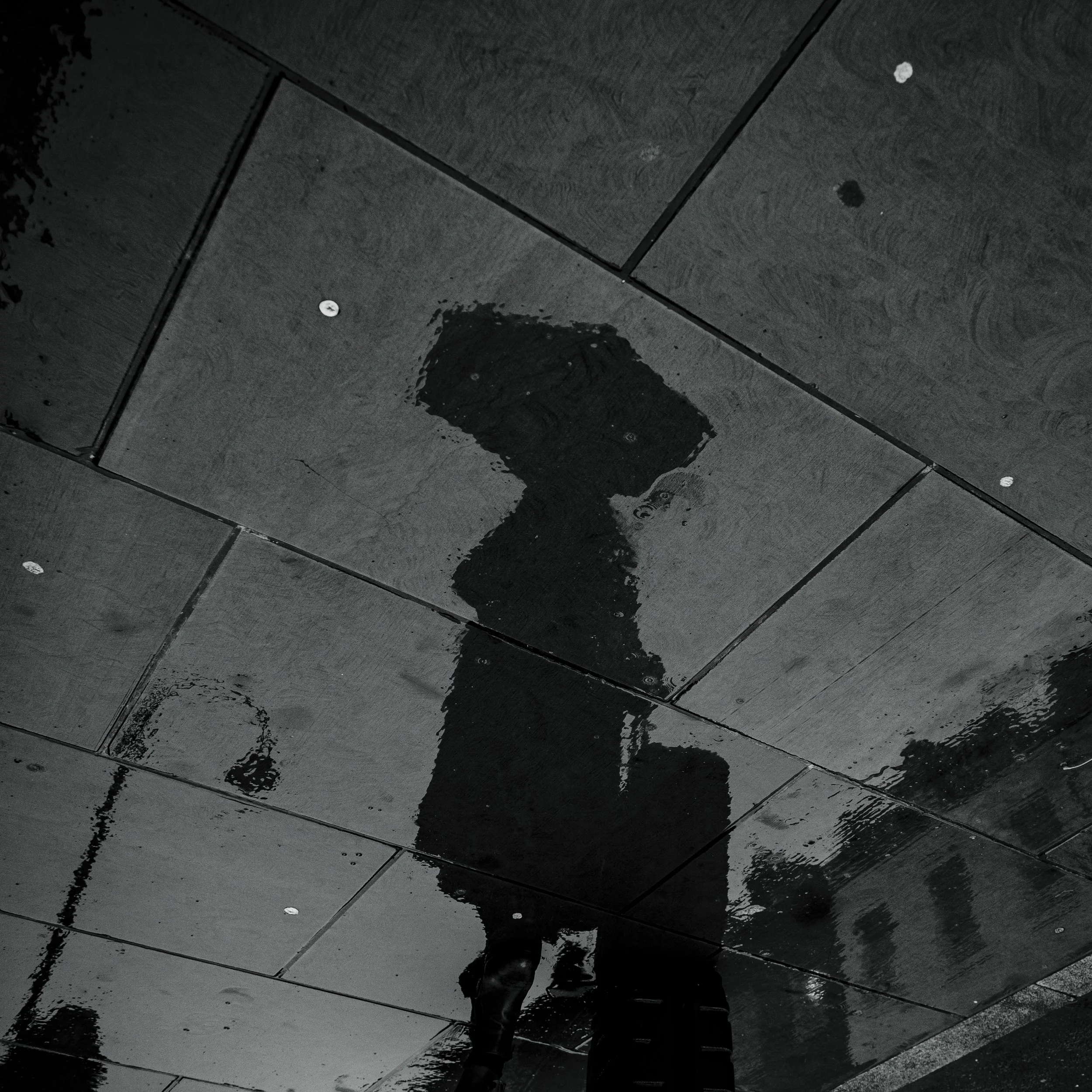 Reflection of a person walking on wet pavement at night, seen in a puddle.