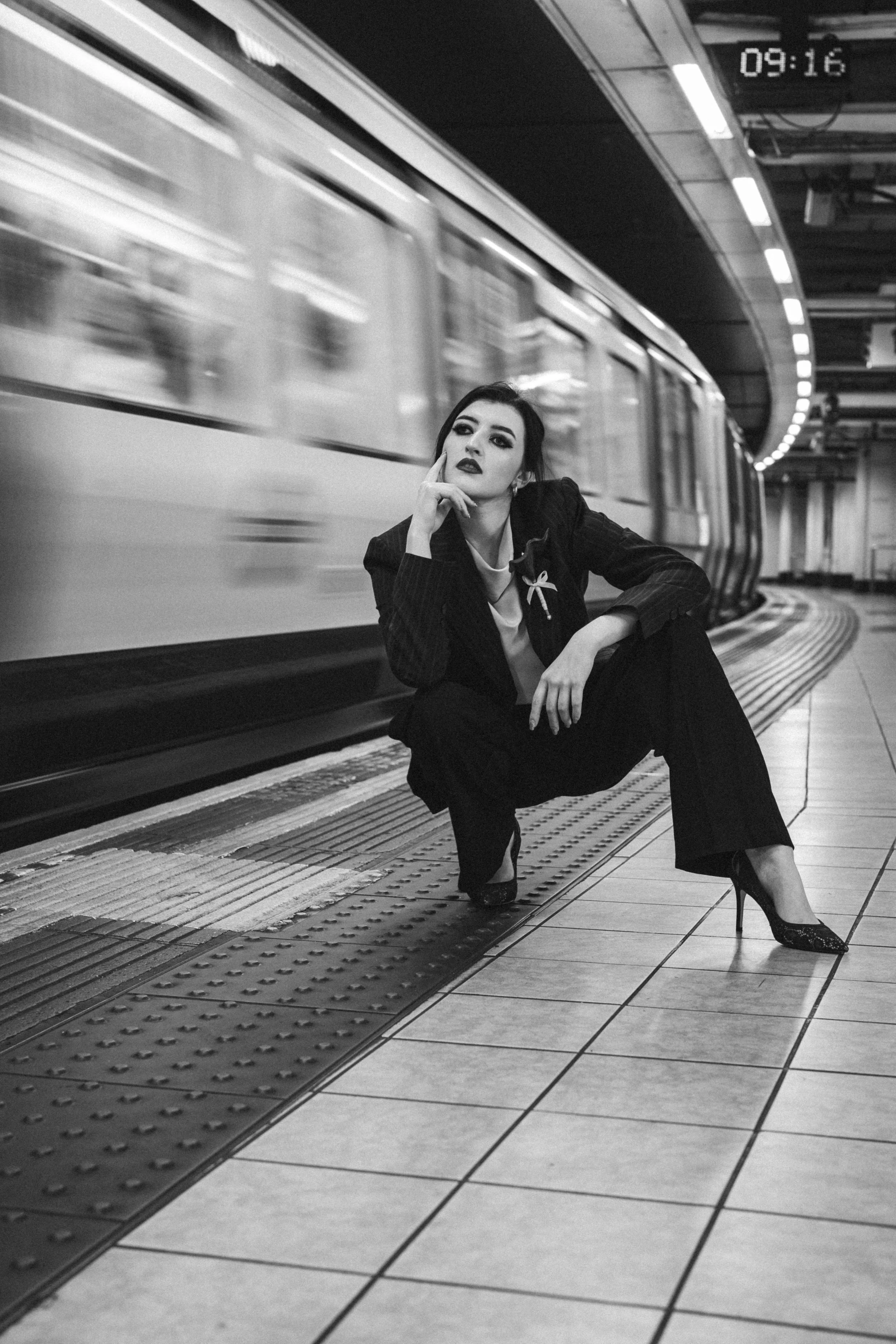 A woman with dark hair and makeup, wearing a striped blazer and high heels, squatting on a subway platform in front of a moving train. The platform has tactile paving, and the timestamp in the top right corner reads 09:16.